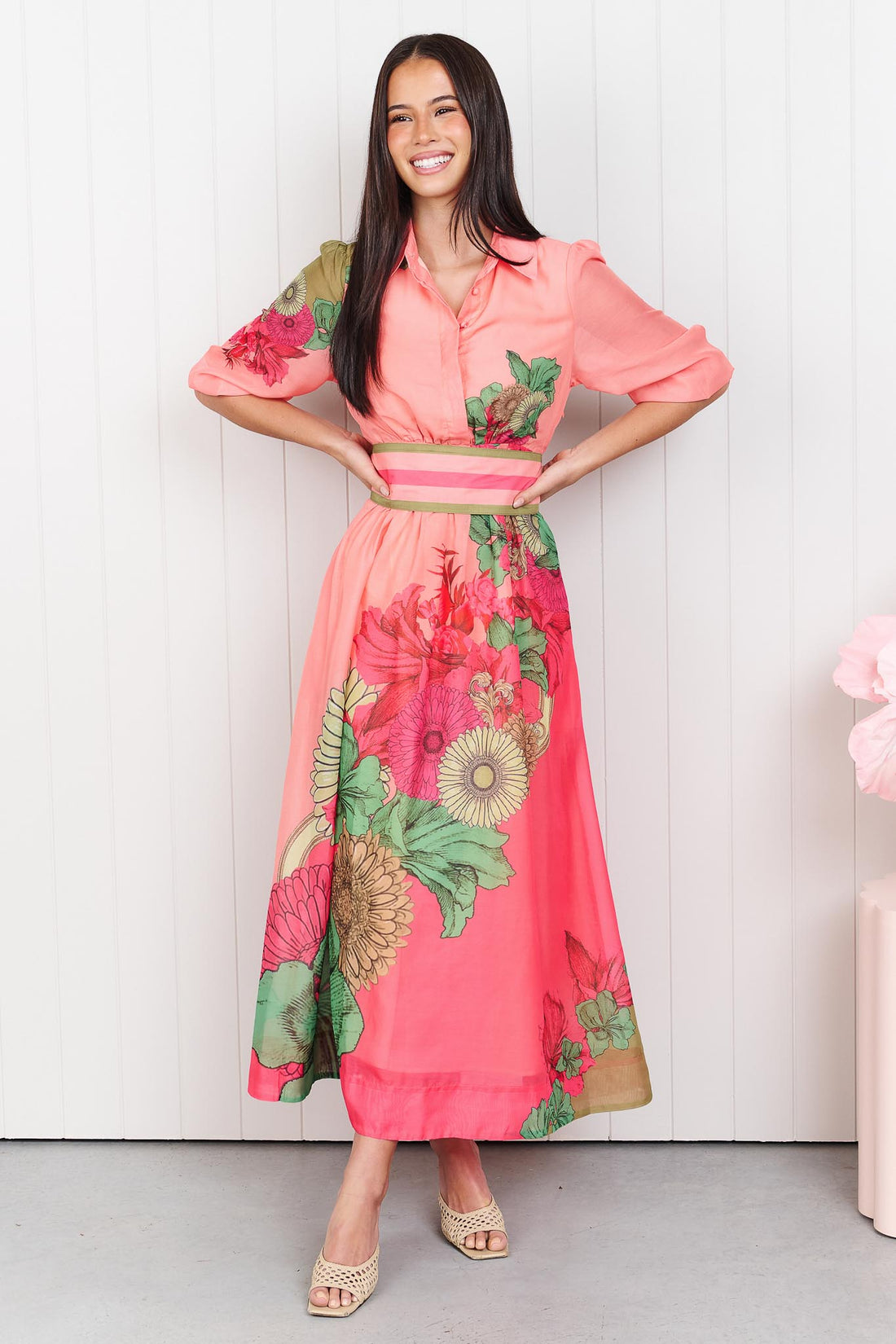 Woman wearing the minnie + MAXXIE Ayla Madison Ave midi event dress in peach and pink with a floral print and stripe waist tie, standing in front of a white panelled wall.