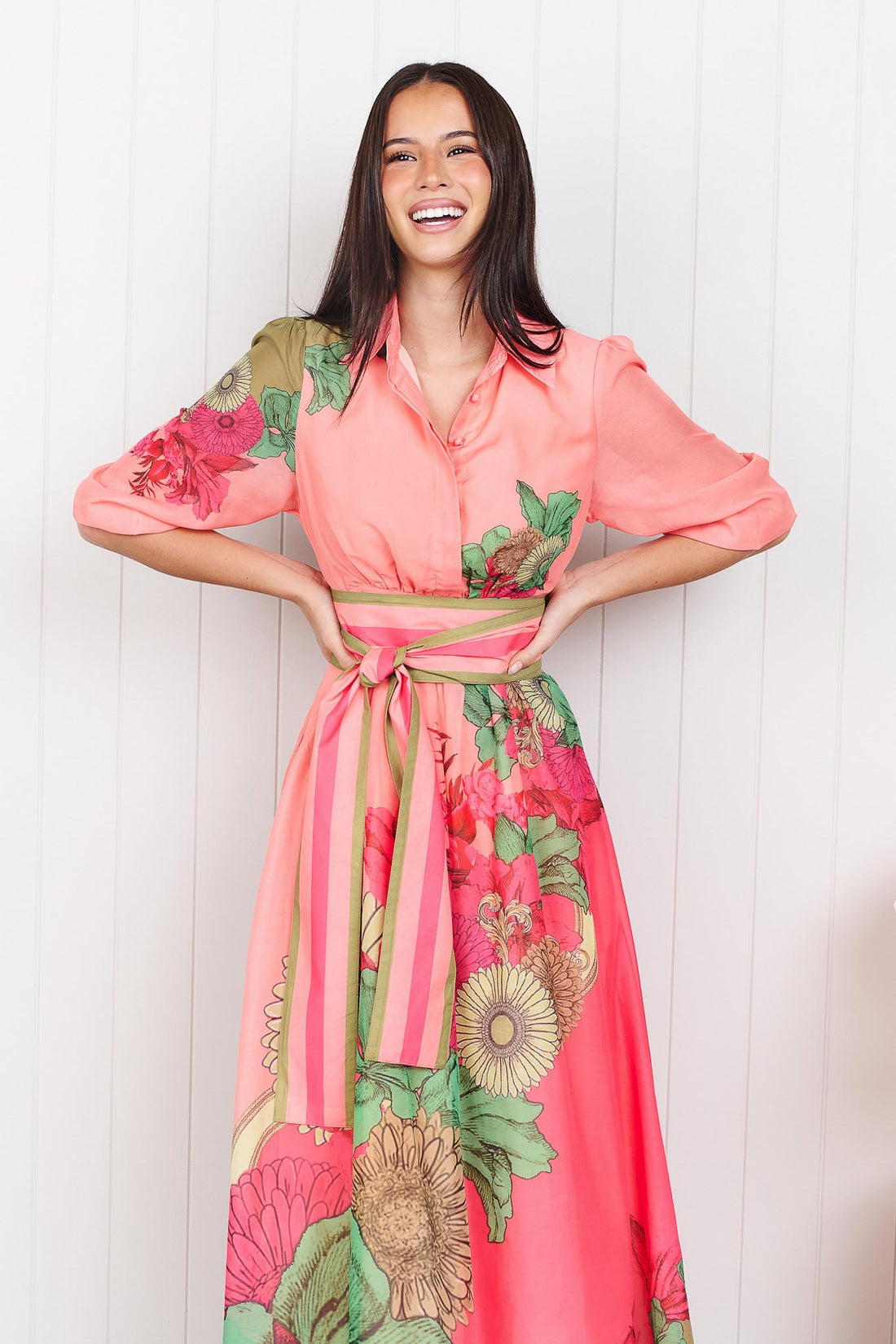 Woman wearing the minnie + MAXXIE Ayla Madison Ave midi event dress in peach and pink with a floral print and stripe waist tie, standing in front of a white panelled wall.