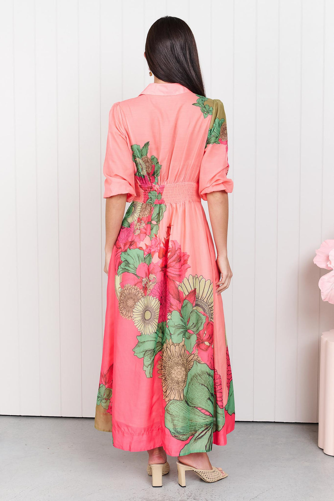 Back view of a woman wearing the minnie + MAXXIE Ayla Madison Ave midi event dress in peach and pink with a floral print, standing in front of a white panelled wall.
