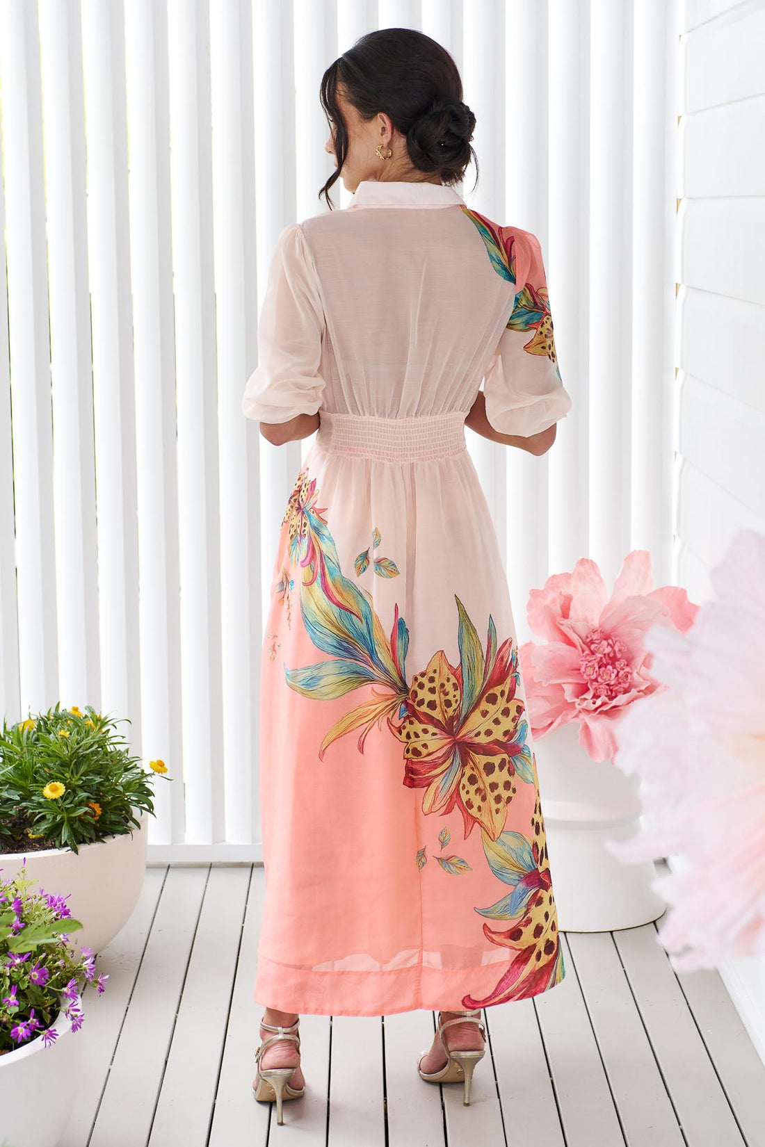 Back view of a woman wearing the minnie + MAXXIE Ayla Havana midi event dress in soft peach and pink with a leopard floral print, standing in front of a white panelled wall.
