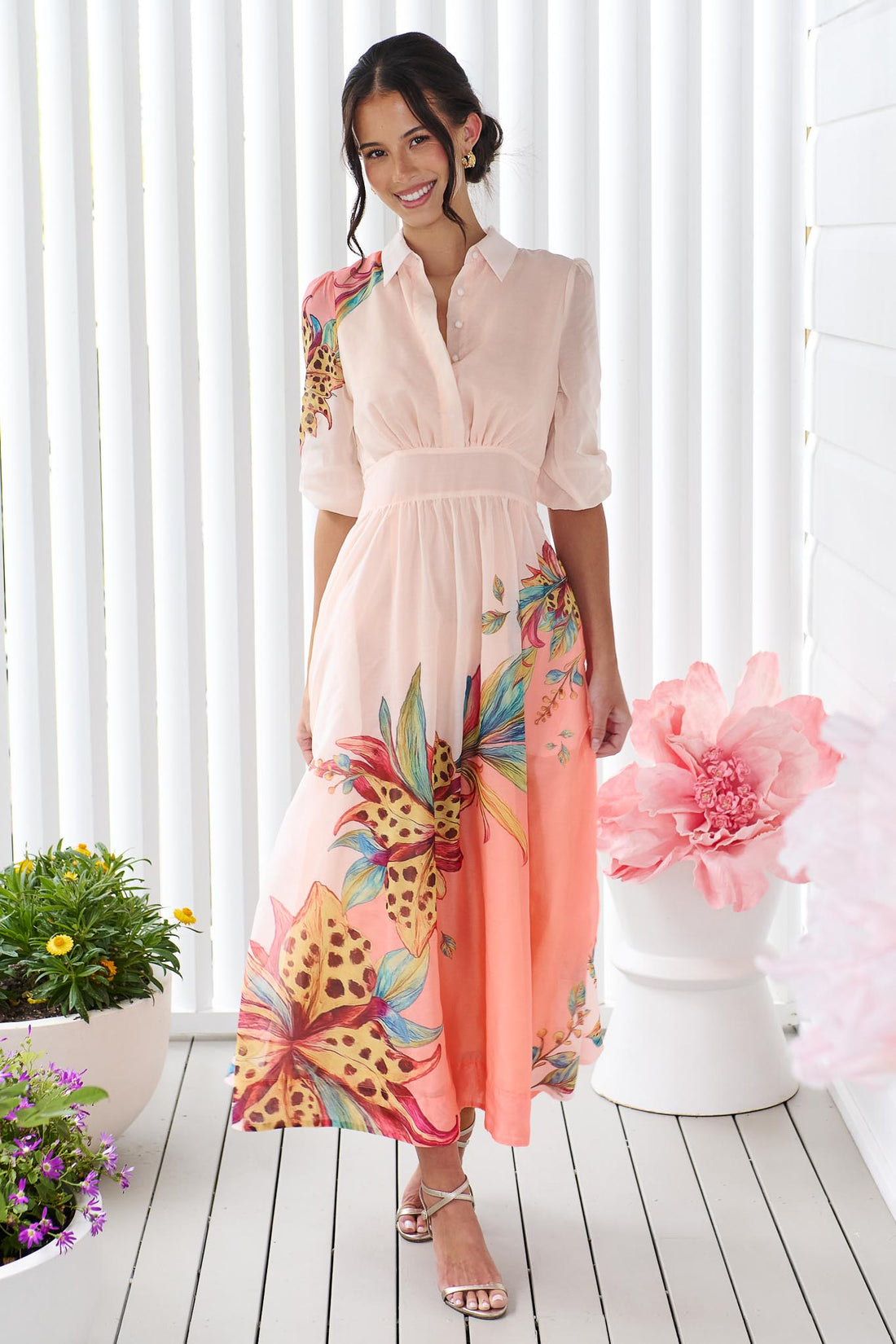 Woman wearing the minnie + MAXXIE Ayla Havana midi event dress in soft peach and pink with a leopard floral print, standing in front of a white panelled wall.