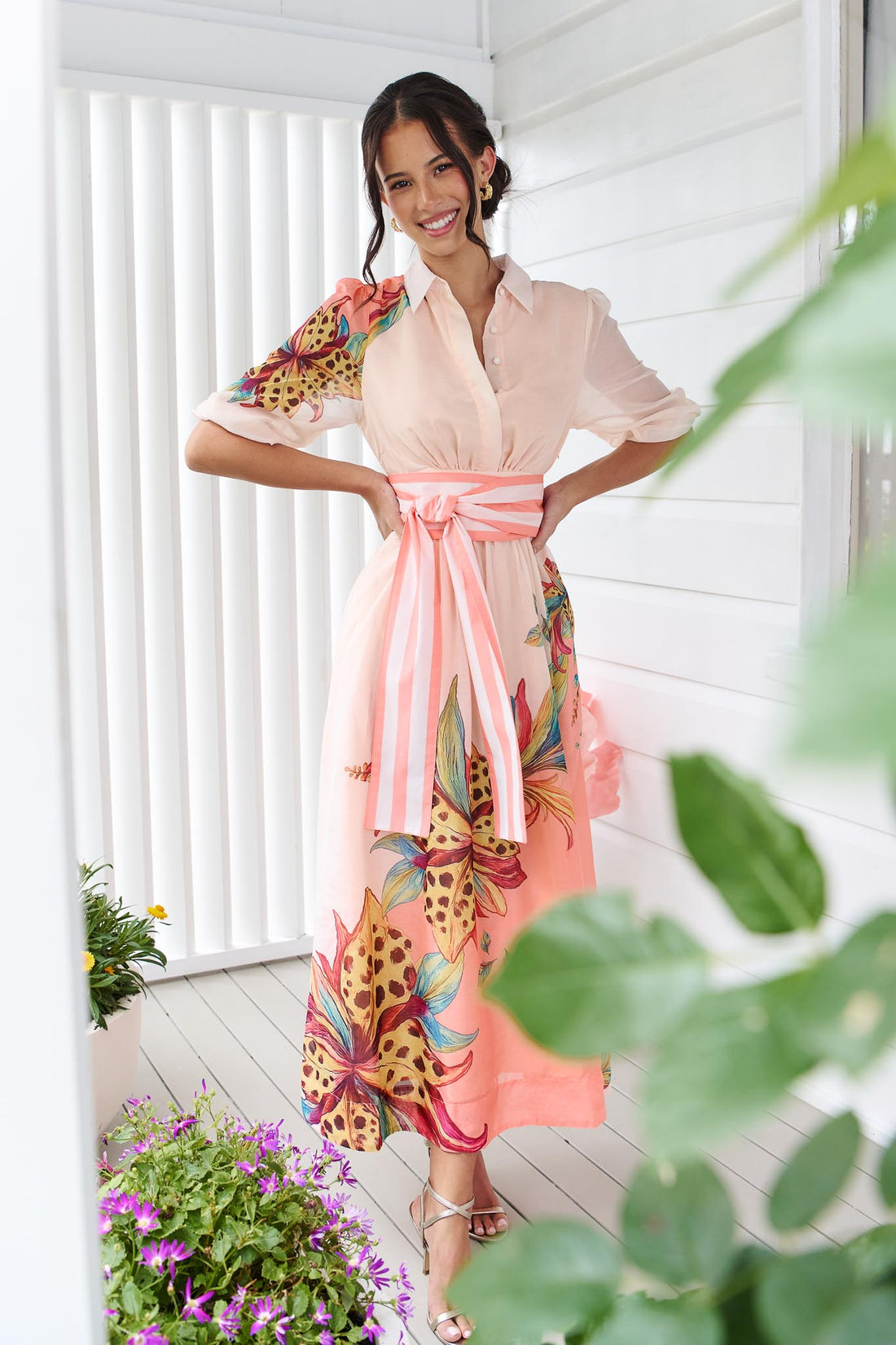 Woman wearing the minnie + MAXXIE Ayla Havana midi event dress in soft peach and pink with a leopard floral print and stripe waist tie, standing in front of a white panelled wall.