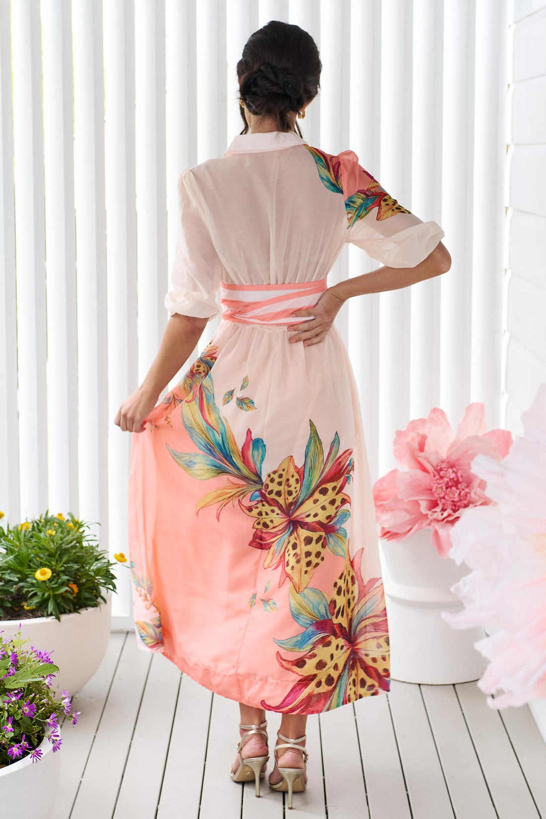 Back view of a woman wearing the minnie + MAXXIE Ayla Havana midi event dress in soft peach and pink with a leopard floral print and stripe waist tie, standing in front of a white panelled wall.