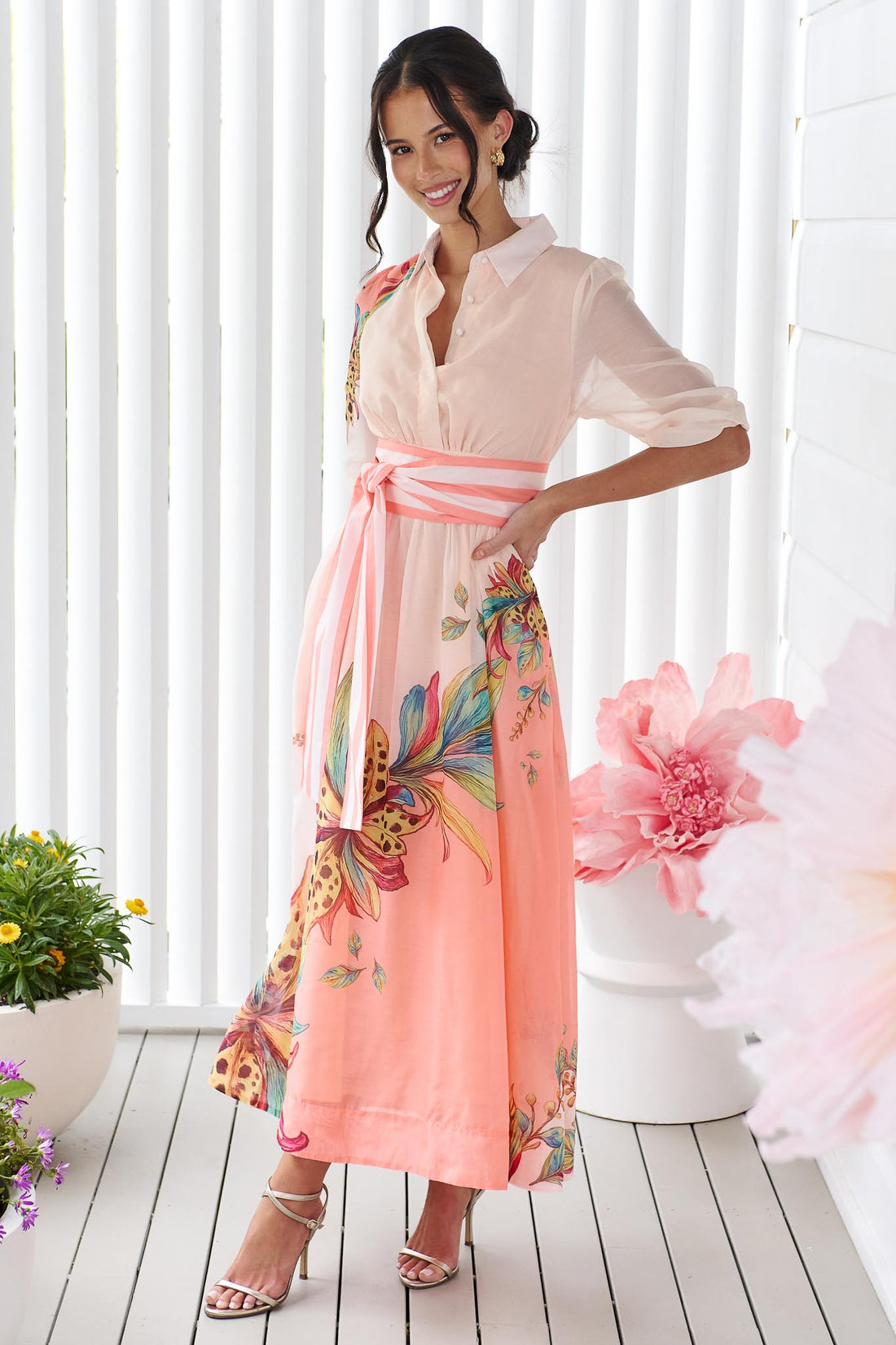 Woman wearing the minnie + MAXXIE Ayla Havana midi event dress in soft peach and pink with a leopard floral print and stripe waist tie, standing in front of a white panelled wall.