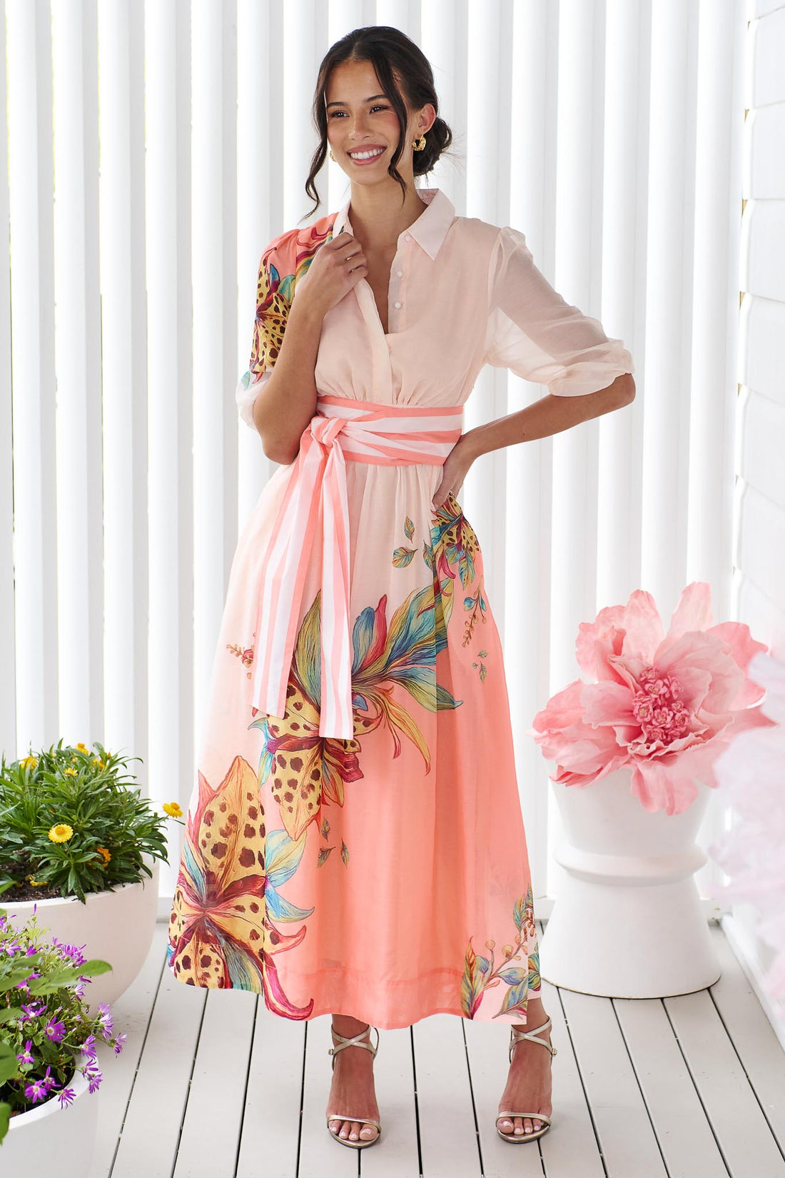 Woman wearing the minnie + MAXXIE Ayla Havana midi event dress in soft peach and pink with a leopard floral print and stripe waist tie, standing in front of a white panelled wall.