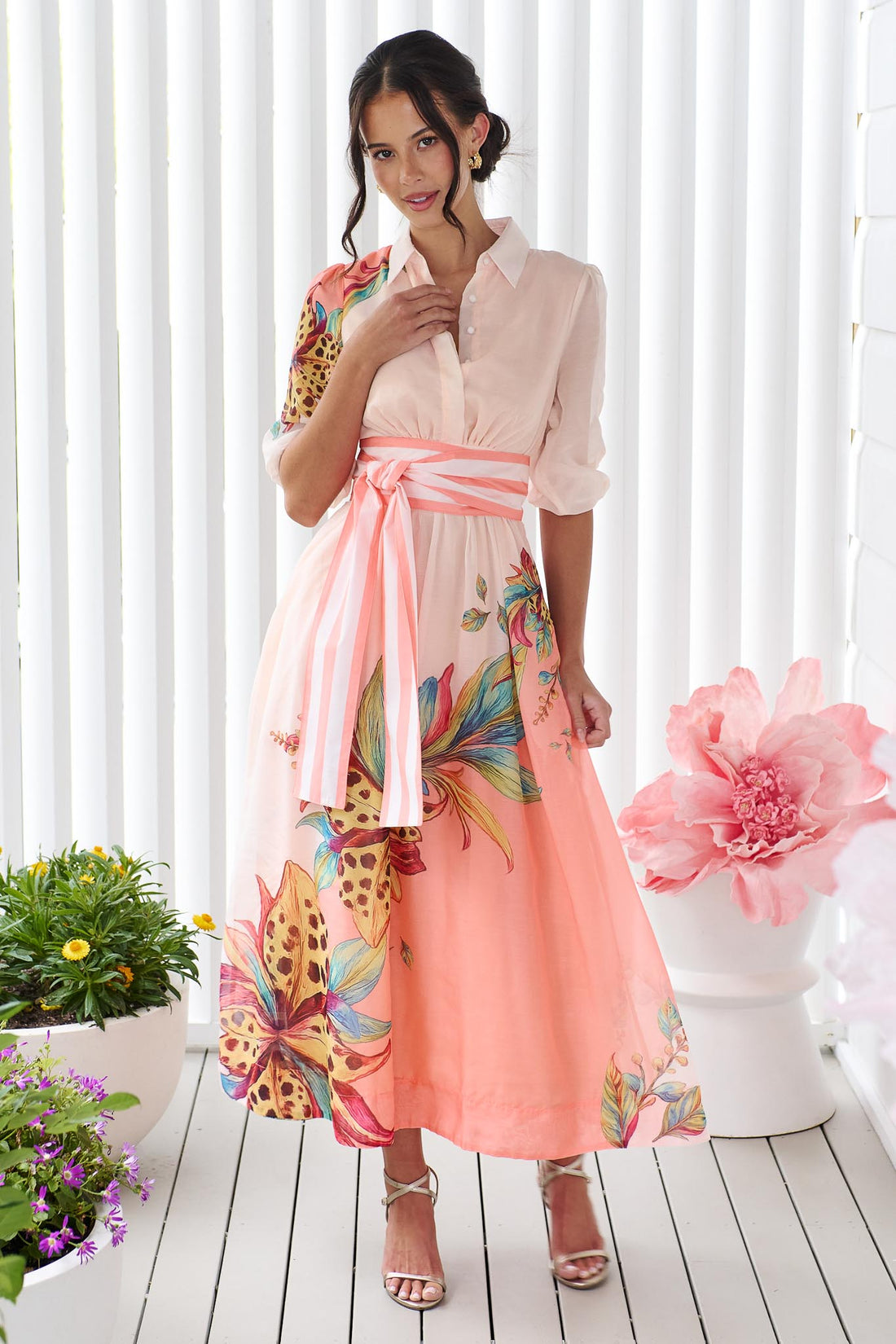 Woman wearing the minnie + MAXXIE Ayla Havana midi event dress in soft peach and pink with a leopard floral print and stripe waist tie, standing in front of a white panelled wall.