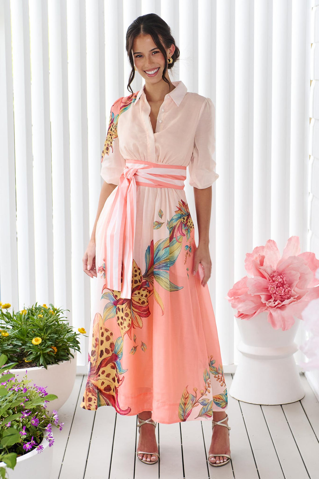 Woman wearing the minnie + MAXXIE Ayla Havana midi event dress in soft peach and pink with a leopard floral print and stripe waist tie, standing in front of a white panelled wall.
