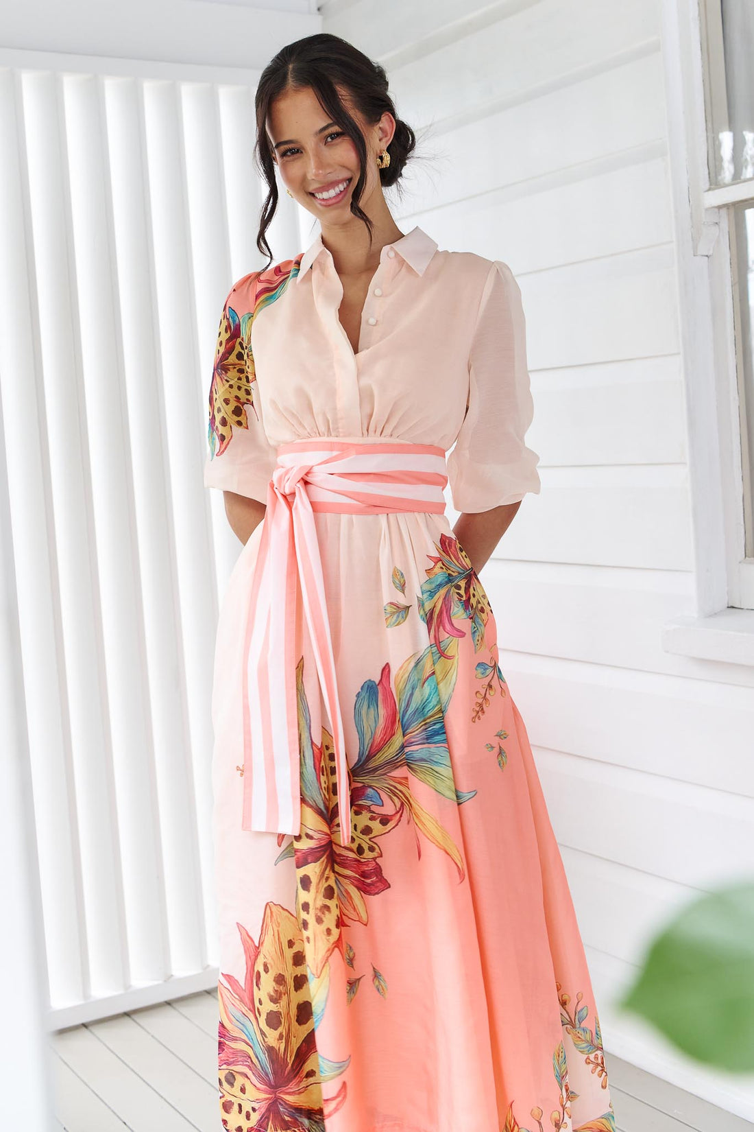 Woman wearing the minnie + MAXXIE Ayla Havana midi event dress in soft peach and pink with a leopard floral print and stripe waist tie, standing in front of a white panelled wall.