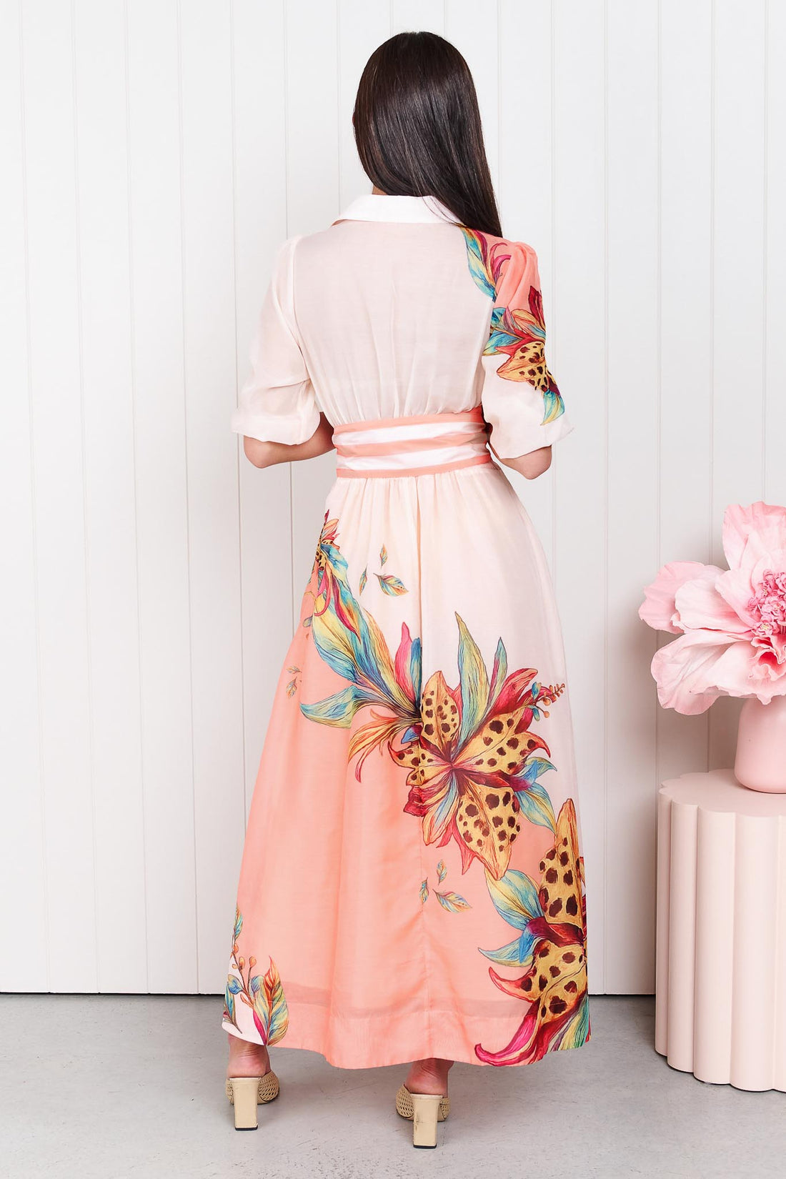 Back view of a woman wearing the minnie + MAXXIE Ayla Havana midi event dress in soft peach and pink with a leopard floral print and stripe waist tie, standing in front of a white panelled wall.