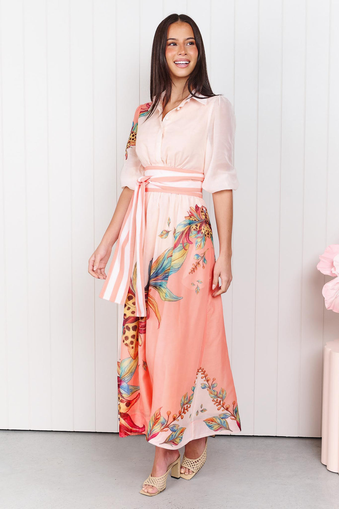 Woman wearing the minnie + MAXXIE Ayla Havana midi event dress in soft peach and pink with a leopard floral print and stripe waist tie, standing in front of a white panelled wall.