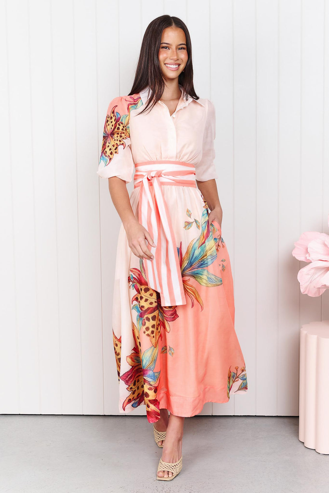 Woman wearing the minnie + MAXXIE Ayla Havana midi event dress in soft peach and pink with a leopard floral print and stripe waist tie, standing in front of a white panelled wall.