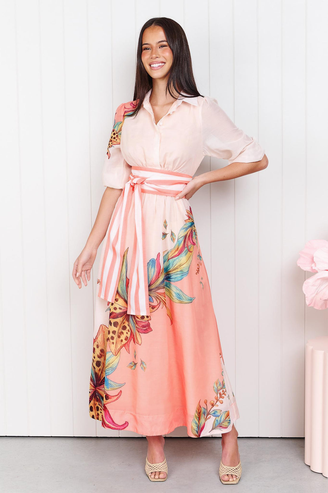 Woman wearing the minnie + MAXXIE Ayla Havana midi event dress in soft peach and pink with a leopard floral print and stripe waist tie, standing in front of a white panelled wall.