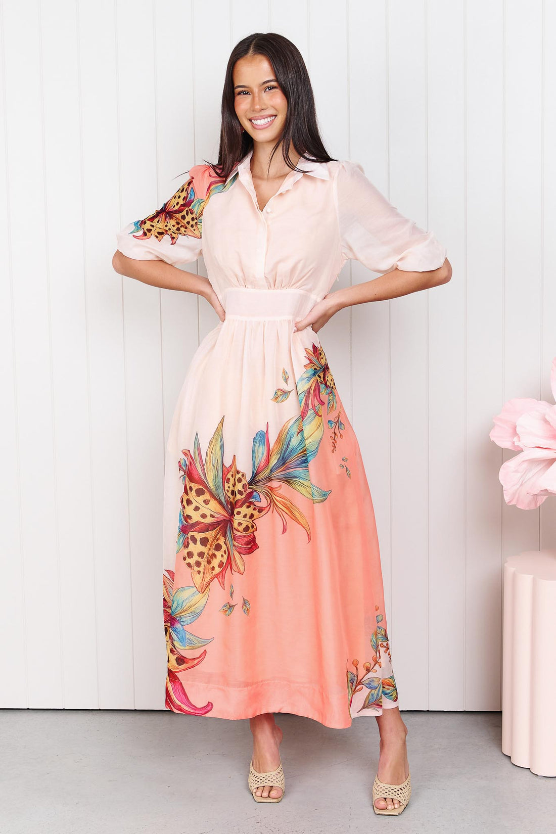 Woman wearing the minnie + MAXXIE Ayla Havana midi event dress in soft peach and pink with a leopard floral print, standing in front of a white panelled wall.