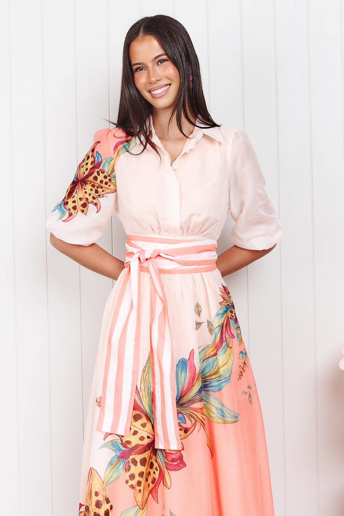 Woman wearing the minnie + MAXXIE Ayla Havana midi event dress in soft peach and pink with a leopard floral print and stripe waist tie, standing in front of a white panelled wall.
