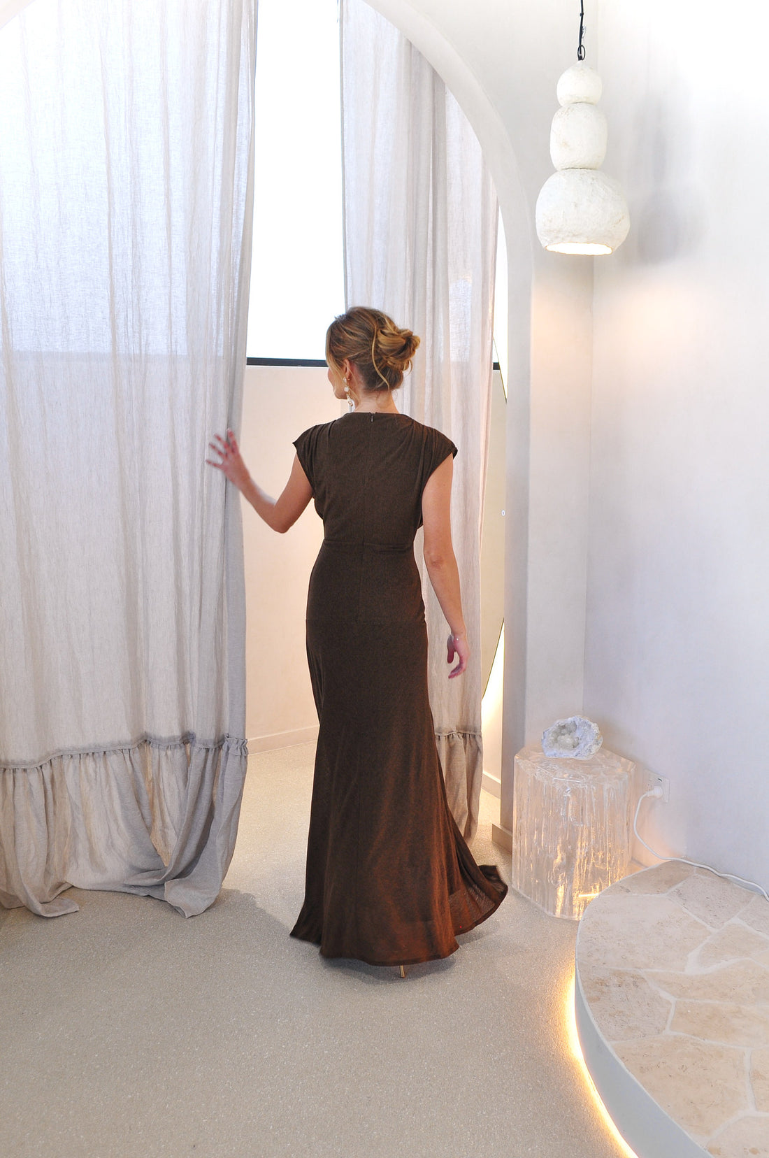 Woman in the cocoa brown Brigitte maxi length event dress standing at the curtains of a fitting room