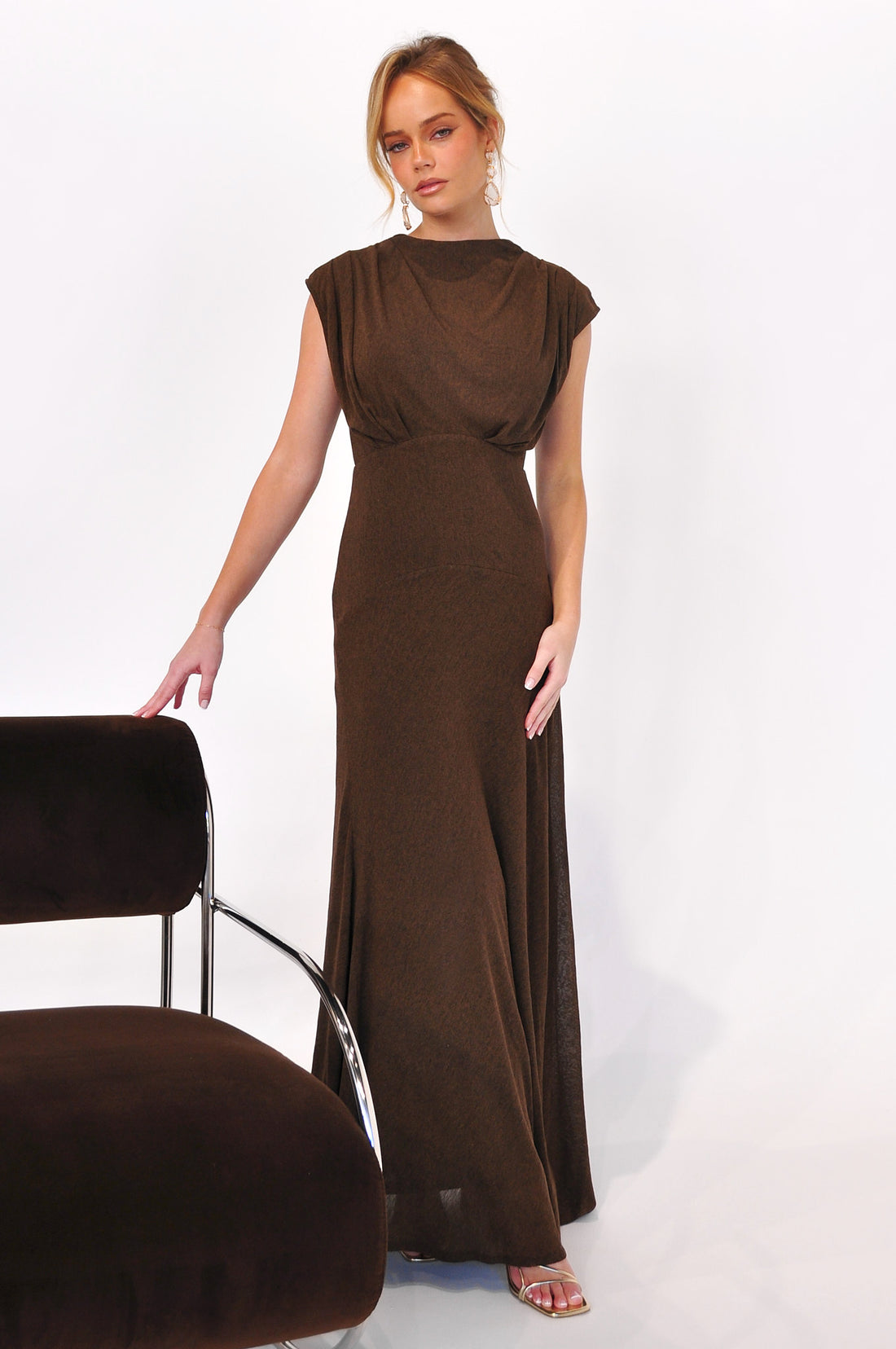 Woman wearing the cocoa brown Brigitte Gown, a maxi length event dress, standing next to a brown velvet chair