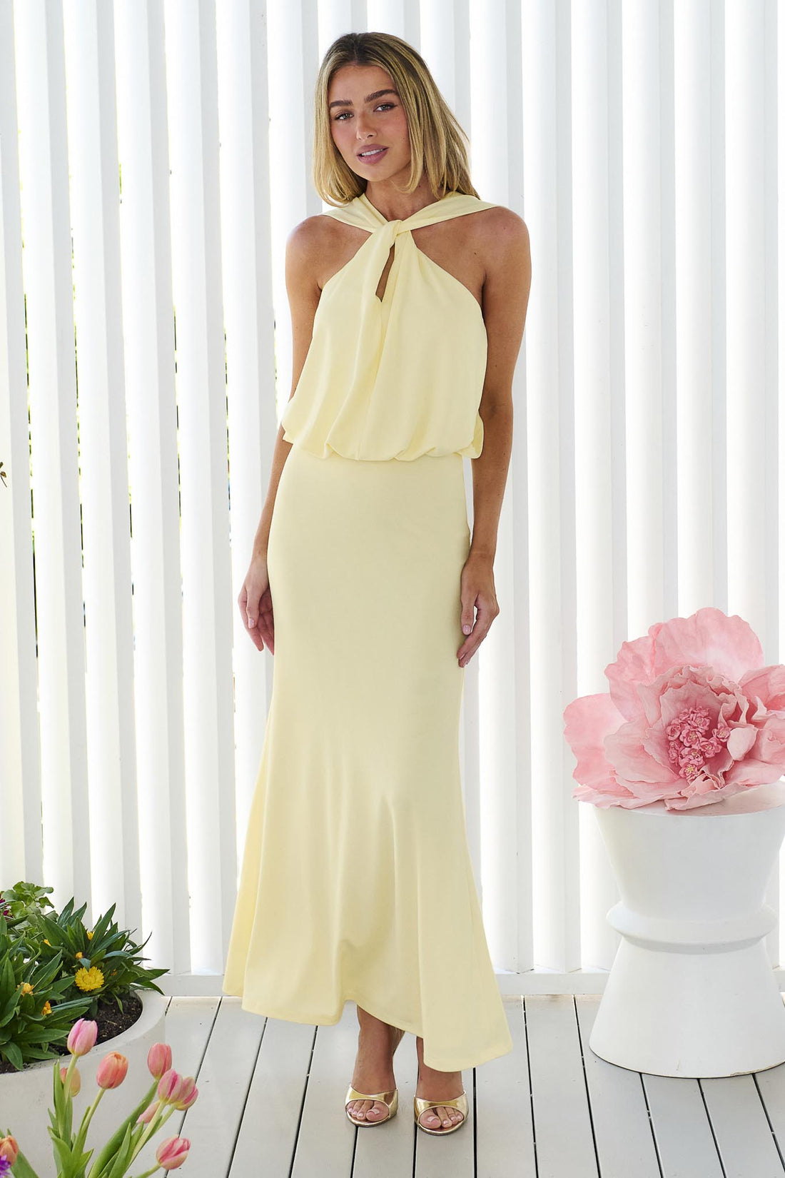 A woman wearing the minnie + MAXXIE Brooklyn halter neck maxi event dress in butter yellow, standing in front of a white panel wall.
