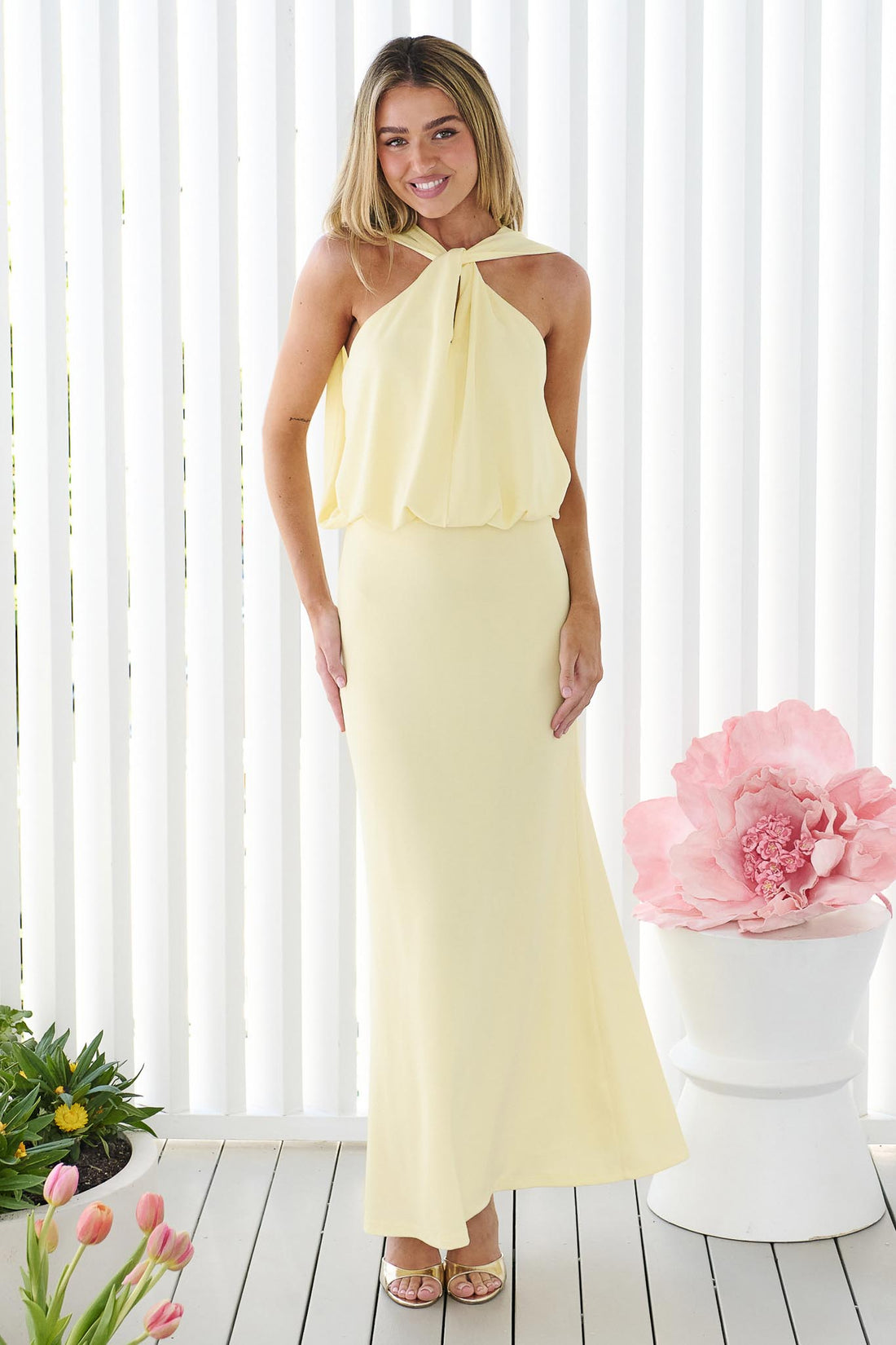 A woman wearing the minnie + MAXXIE Brooklyn halter neck maxi event dress in butter yellow, standing in front of a white panel wall.