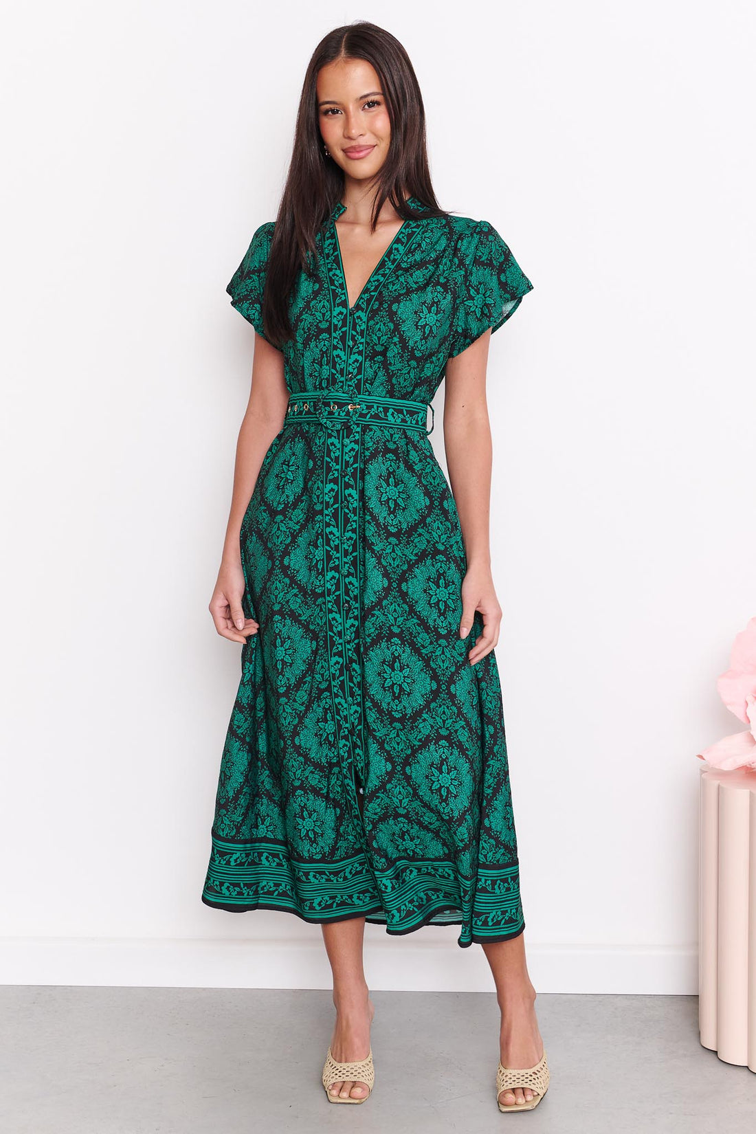 A woman wearing the minnie + MAXXIE green and black print Caitlin midi dress with short flutter sleeves, a V-neckline, and a matching belt, standing against a white wall.