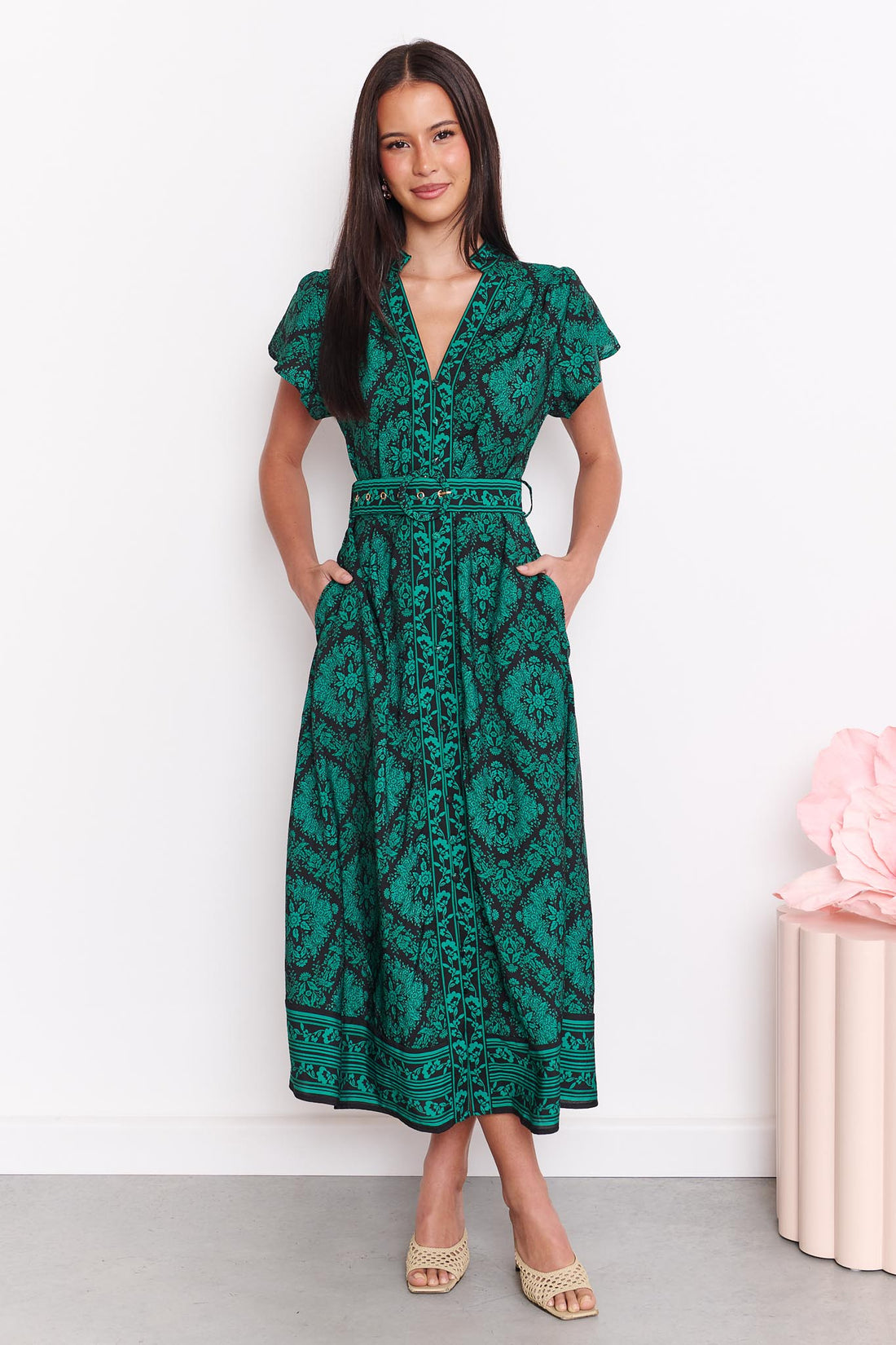 A woman wearing the minnie + MAXXIE green and black print Caitlin midi dress with short flutter sleeves, a V-neckline, side pockets and a matching belt, standing against a white wall.