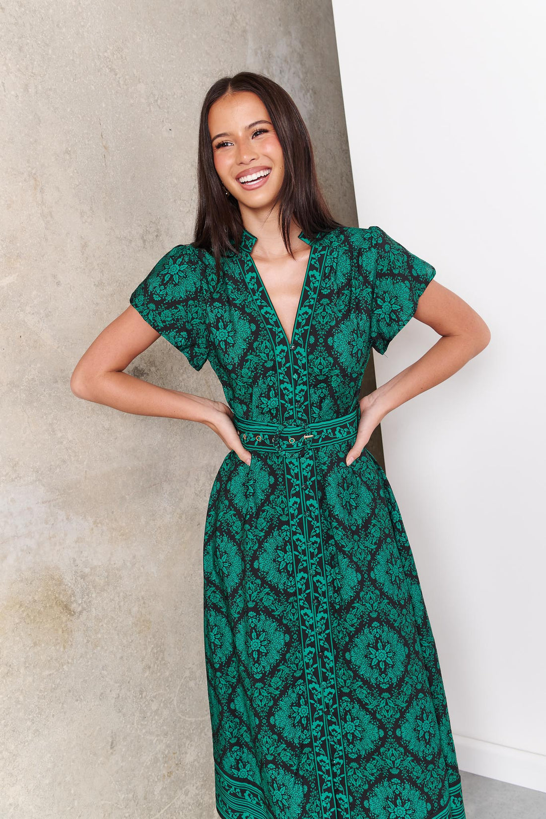 A woman wearing the minnie + MAXXIE green and black print Caitlin midi dress with short flutter sleeves, a V-neckline, and a matching belt, standing against a concrete wall.