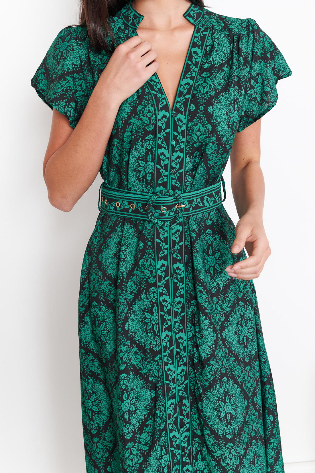 Detail of a woman wearing the minnie + MAXXIE green and black print Caitlin midi dress with short flutter sleeves, a V-neckline, and a matching belt, standing against a white wall.