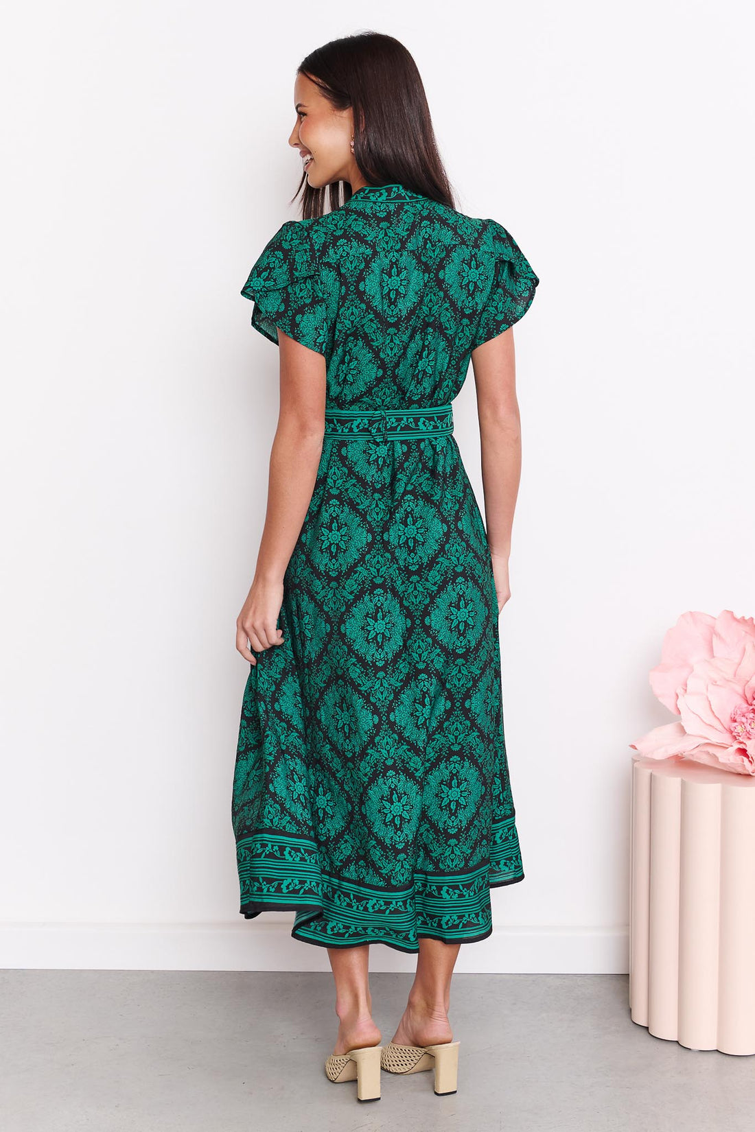 Back view of a woman wearing the minnie + MAXXIE green and black print Caitlin midi dress with short flutter sleeves, a V-neckline, and a matching belt, standing against a white wall.