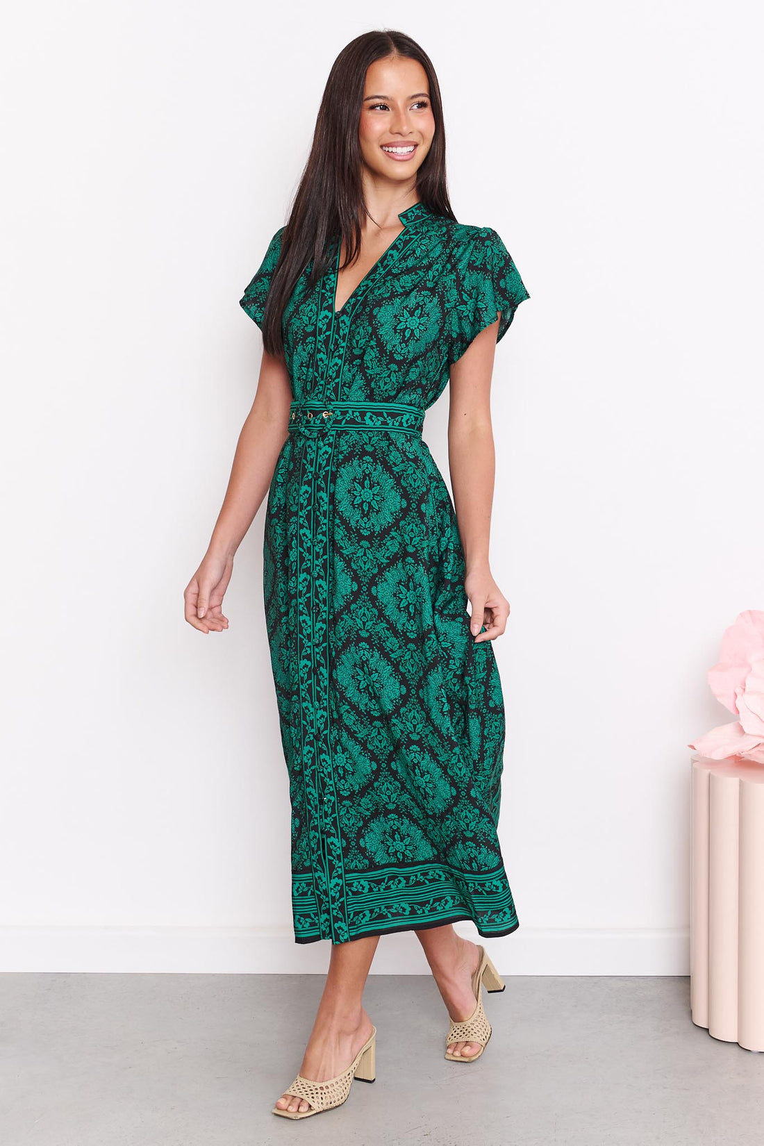 A woman wearing the minnie + MAXXIE green and black print Caitlin midi dress with short flutter sleeves, a V-neckline, and a matching belt, standing against a white wall.