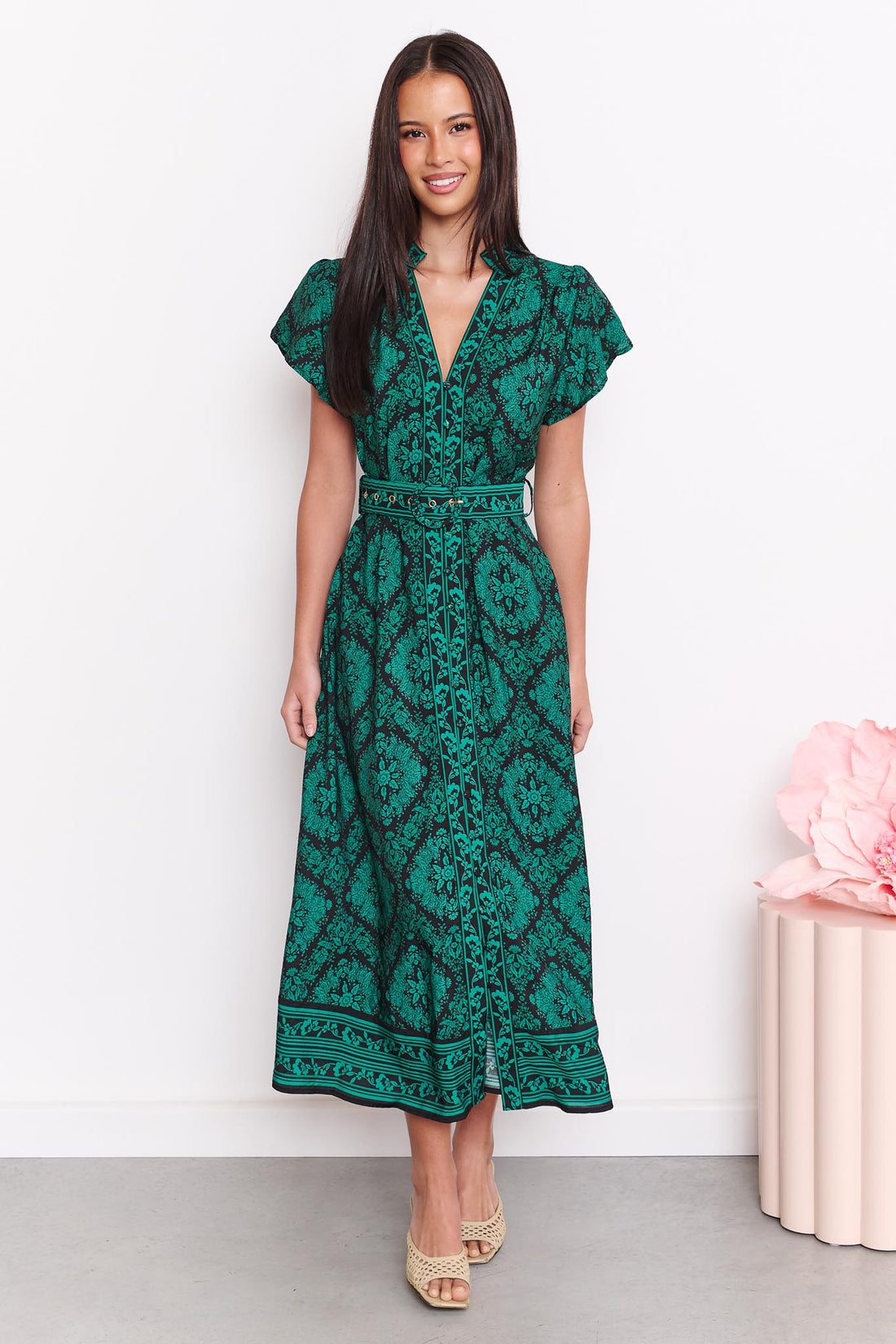 A woman wearing the minnie + MAXXIE green and black print Caitlin midi dress with short flutter sleeves, a V-neckline, and a matching belt, standing against a white wall.