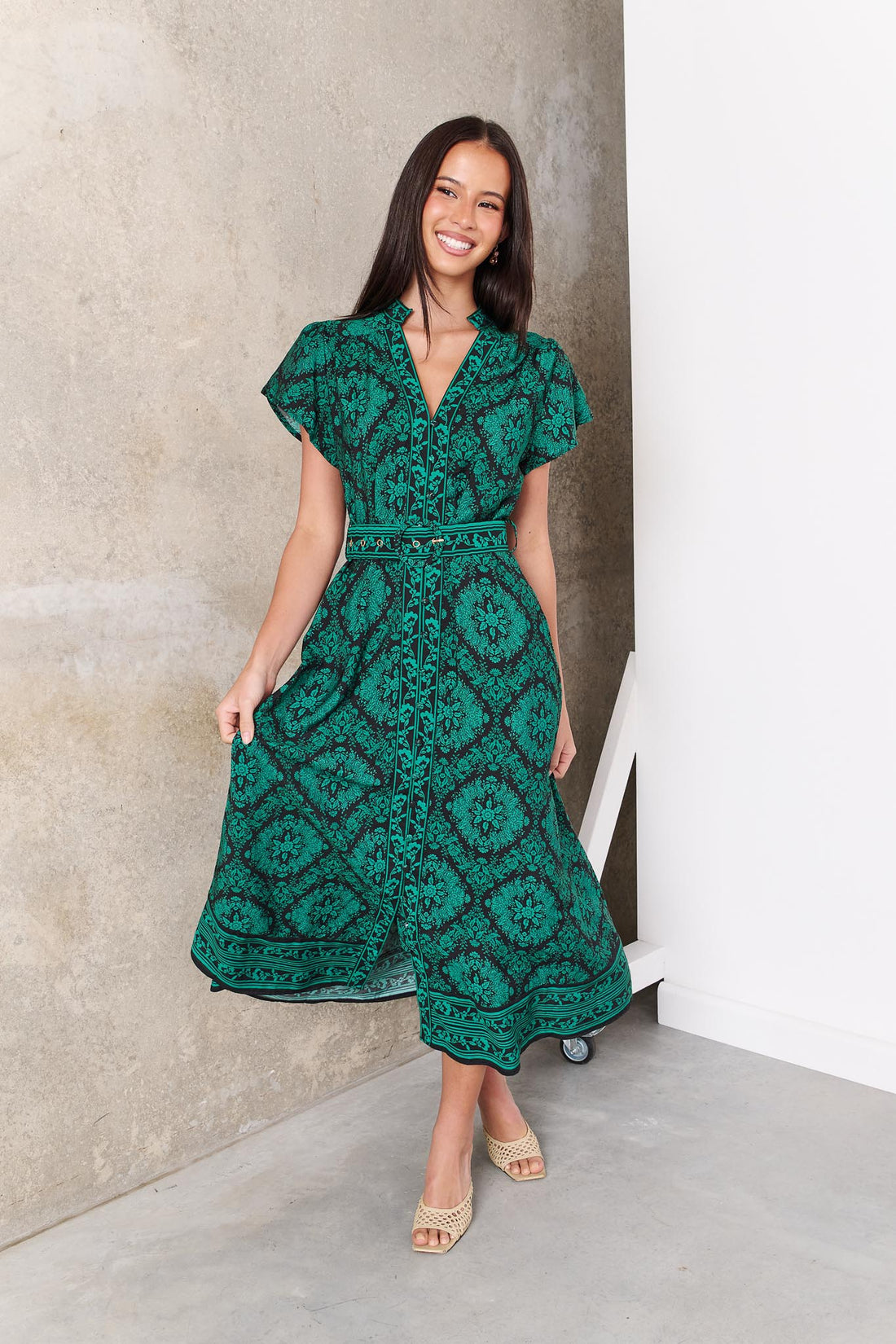 A woman wearing the minnie + MAXXIE green and black print Caitlin midi dress with short flutter sleeves, a V-neckline, and a matching belt, standing against a concrete wall.