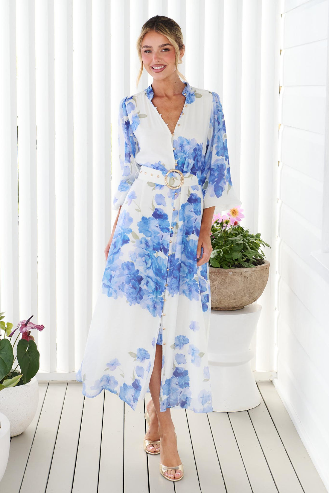 A woman wearing the minnie + MAXXIE Carey white long-sleeve midi dress with a vibrant blue floral print and a matching belt, standing in front of a white panelled wall.