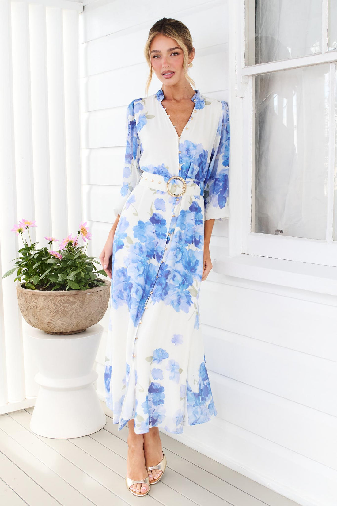 A woman wearing the minnie + MAXXIE Carey white long-sleeve midi dress with a vibrant blue floral print and a matching belt, standing in front of a white window.