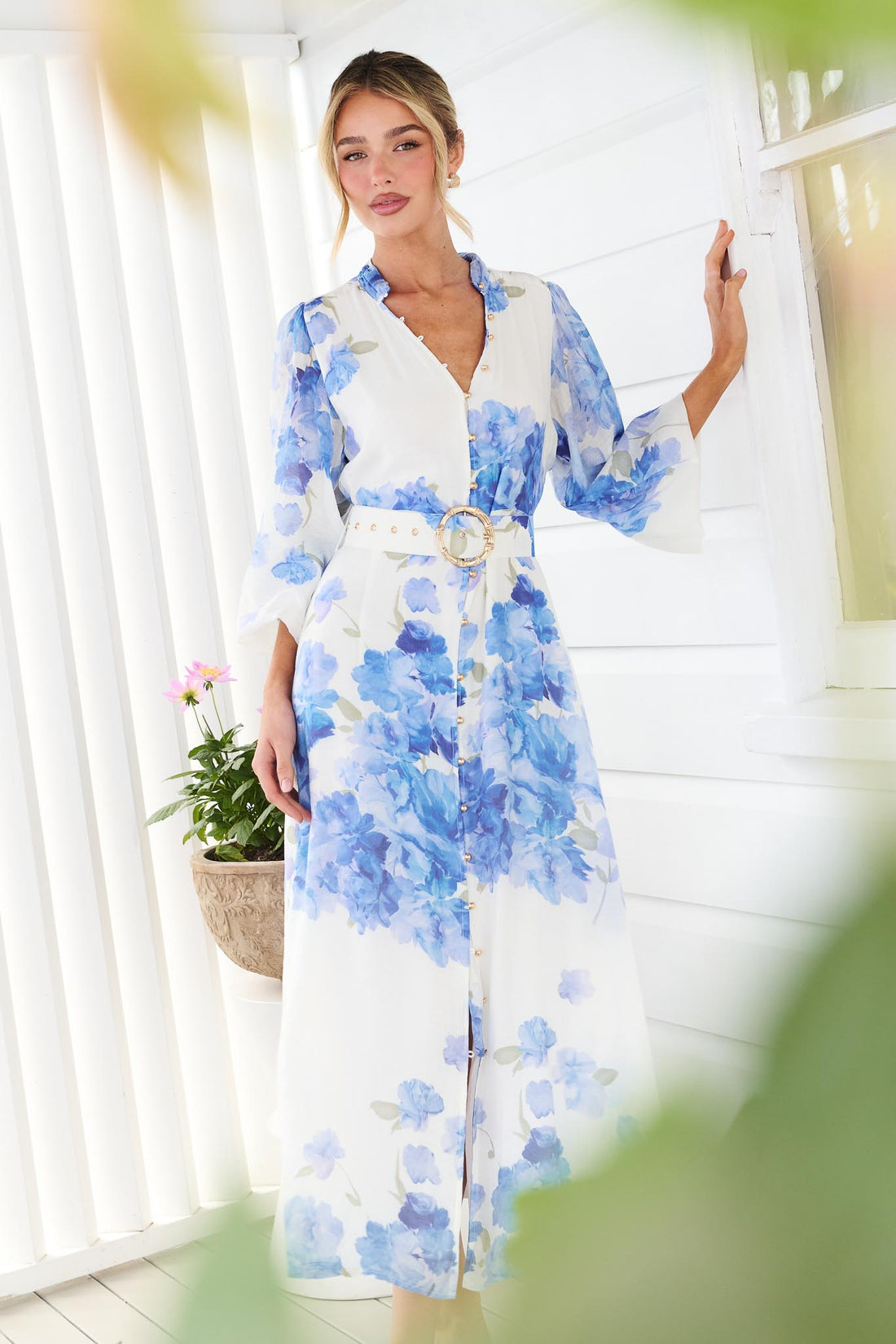 A woman wearing the minnie + MAXXIE Carey white long-sleeve midi dress with a vibrant blue floral print and a matching belt, standing in front of a white window.