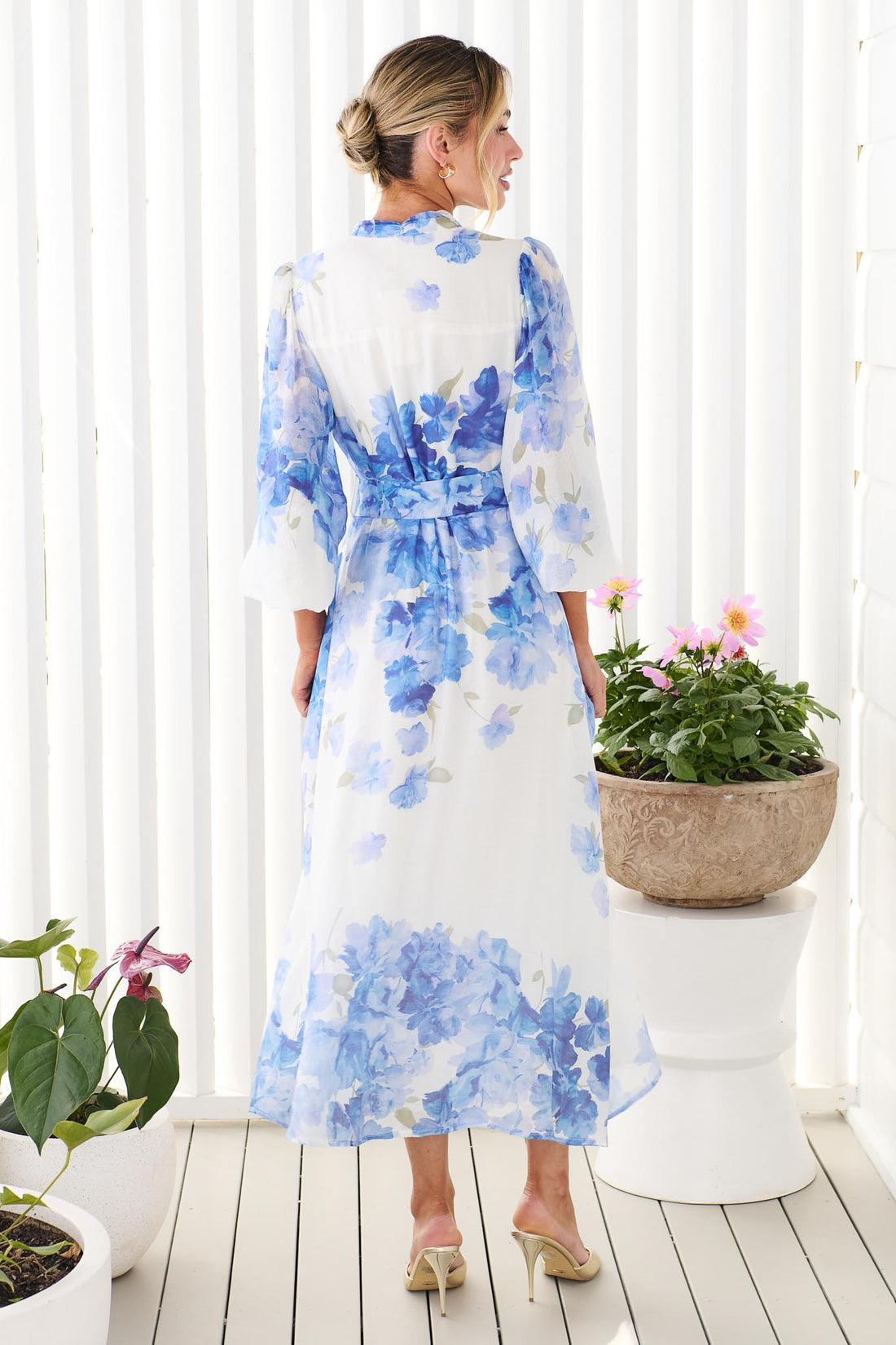 Back view of a woman wearing the minnie + MAXXIE Carey white long-sleeve midi dress with a vibrant blue floral print and a matching belt, standing in front of a white panelled wall.