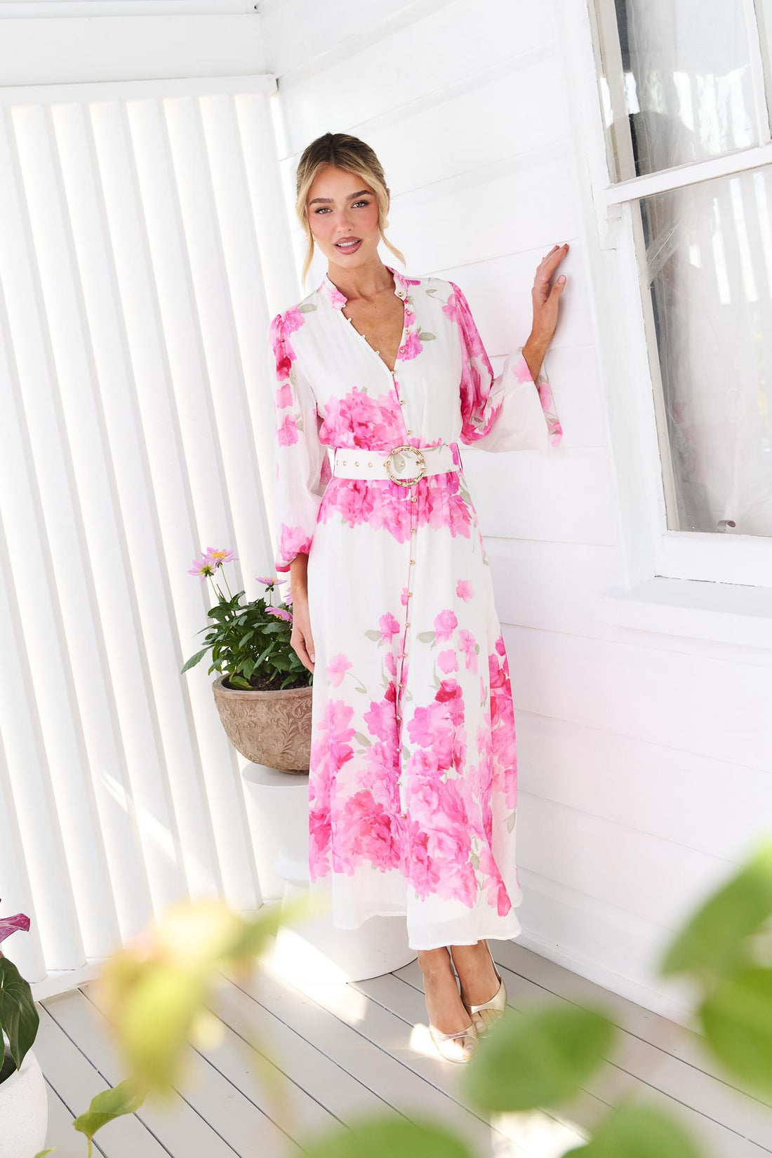 A woman wearing the minnie + MAXXIE Carey white long-sleeve midi dress with a vibrant pink floral print and a matching belt, standing in front of a white window.
