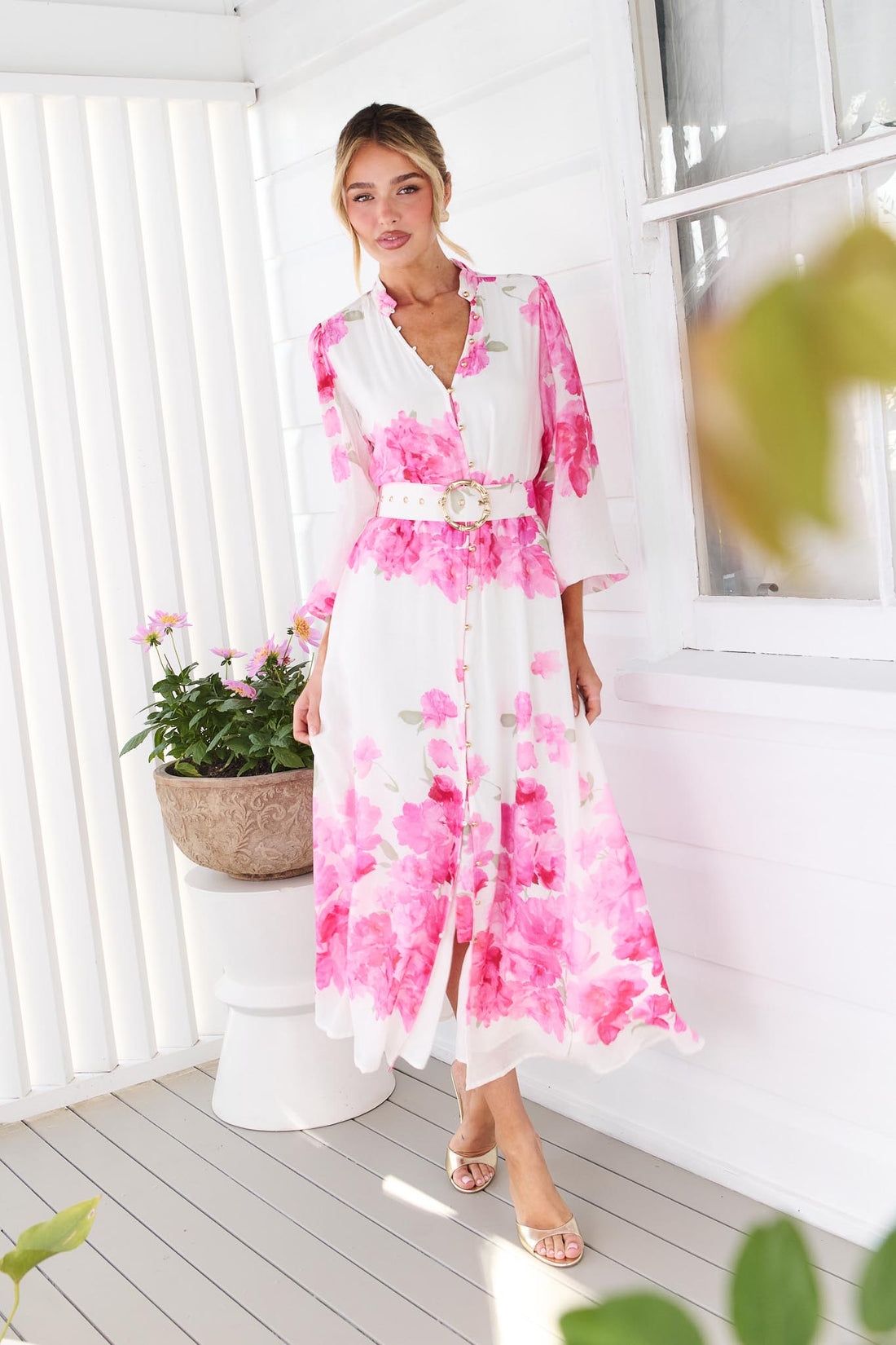 A woman wearing the minnie + MAXXIE Carey white long-sleeve midi dress with a vibrant pink floral print and a matching belt, standing in front of a white window.