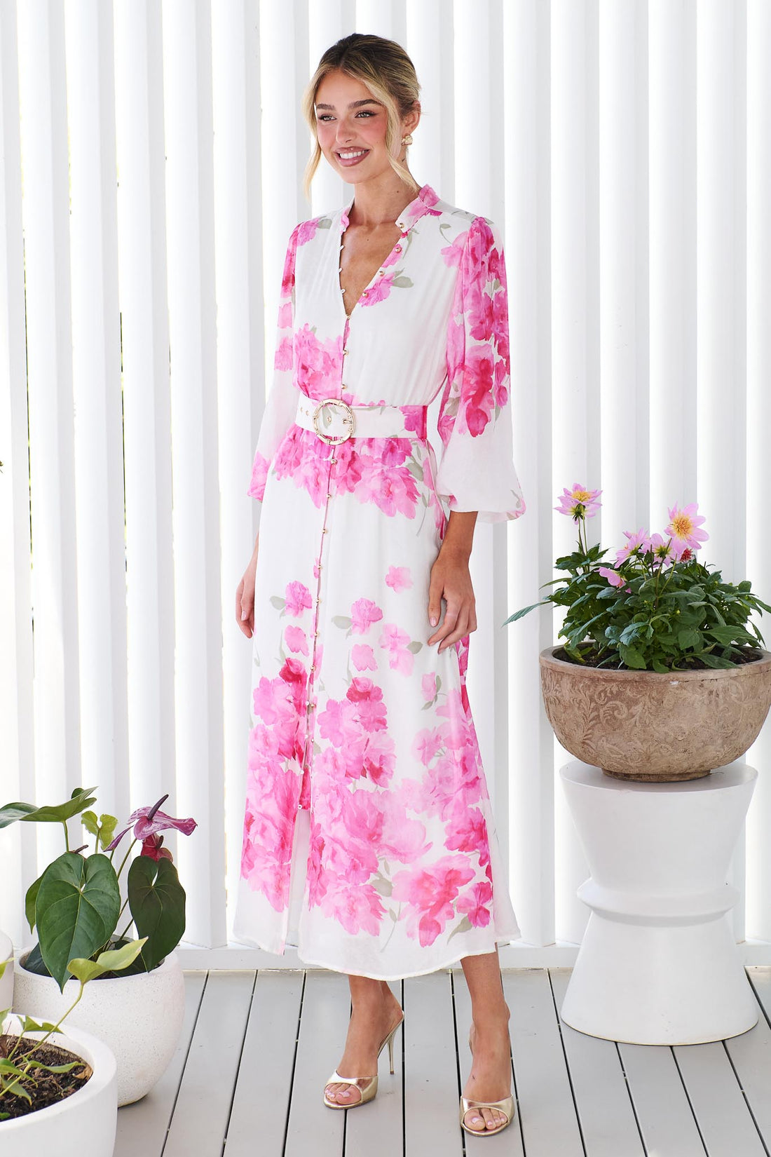 Side view of a woman wearing the minnie + MAXXIE Carey white long-sleeve midi dress with a vibrant pink floral print and a matching belt, standing in front of a white panelled wall.