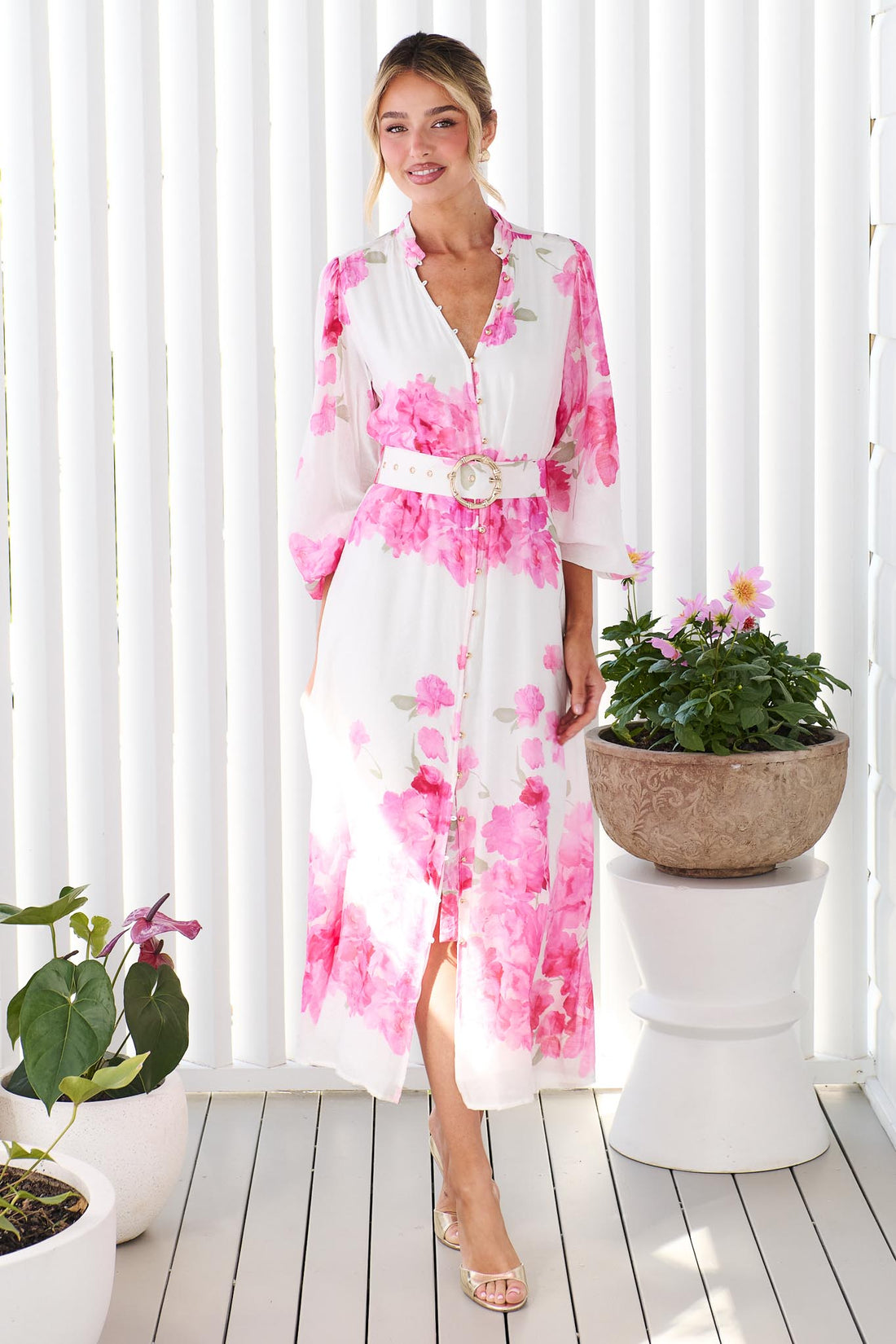 A woman wearing the minnie + MAXXIE Carey white long-sleeve midi dress with a vibrant pink floral print and a matching belt, standing in front of a white panelled wall.