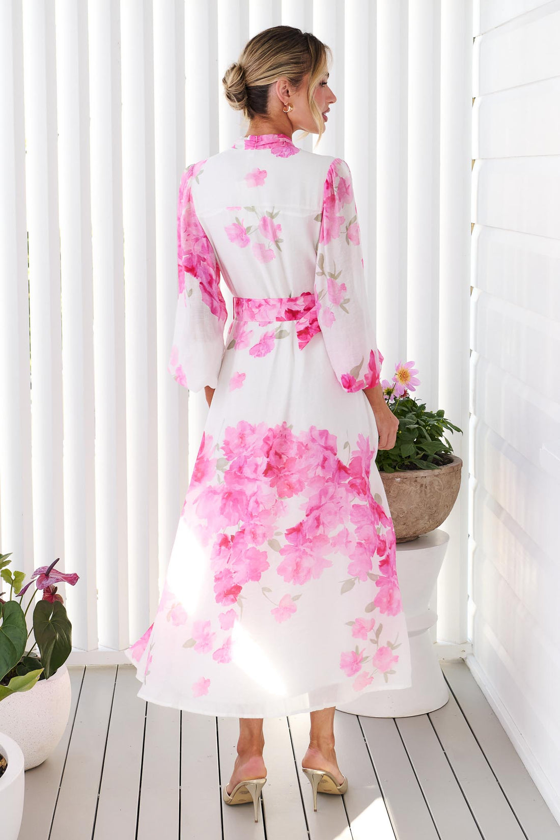 Back view of a woman wearing the minnie + MAXXIE Carey white long-sleeve midi dress with a vibrant pink floral print and a matching belt, standing in front of a white panelled wall.