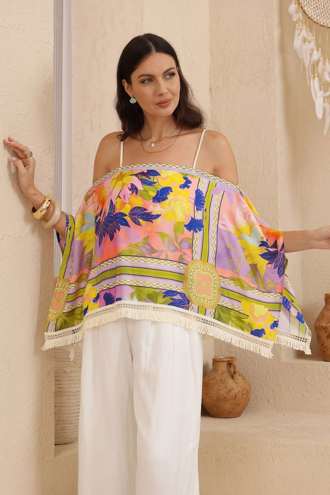 A woman models the minnie + MAXXIE Catalina Resort Top in a vibrant, multicoloured floral print with fringe trim, worn over her shoulders as an off-the-shoulder top, paired with white trousers in a warm, rustic indoor setting.