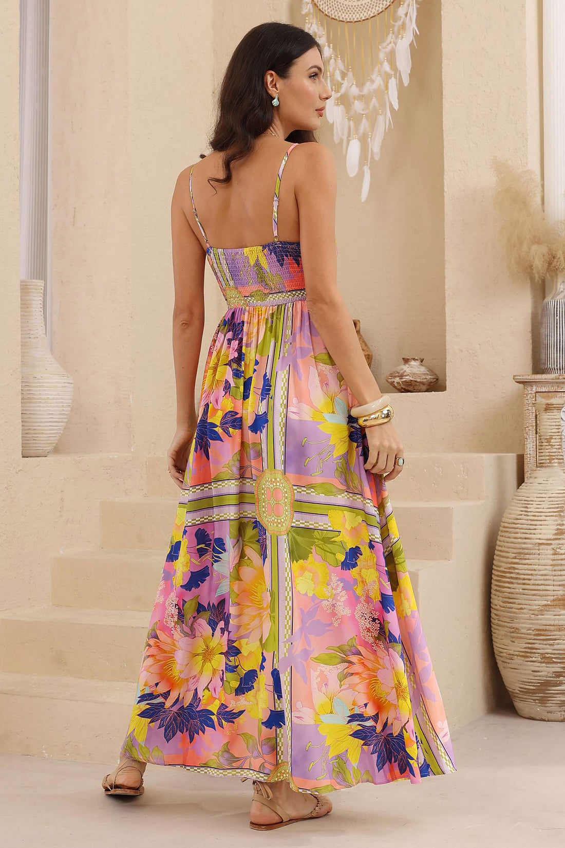 Back view of a woman models the minnie + MAXXIE Catalina maxi dress in a vibrant, multicoloured floral print with a strapless neckline, standing against a textured beige wall next to a white dreamcatcher.