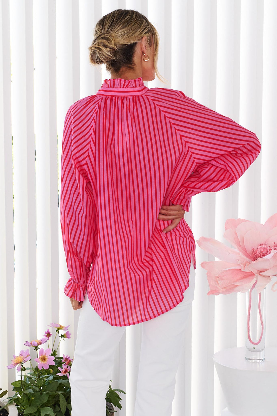 Back view of a woman wearing the vibrant pink and red stripe minnie + MAXXIE Cece Shirt with white pants.