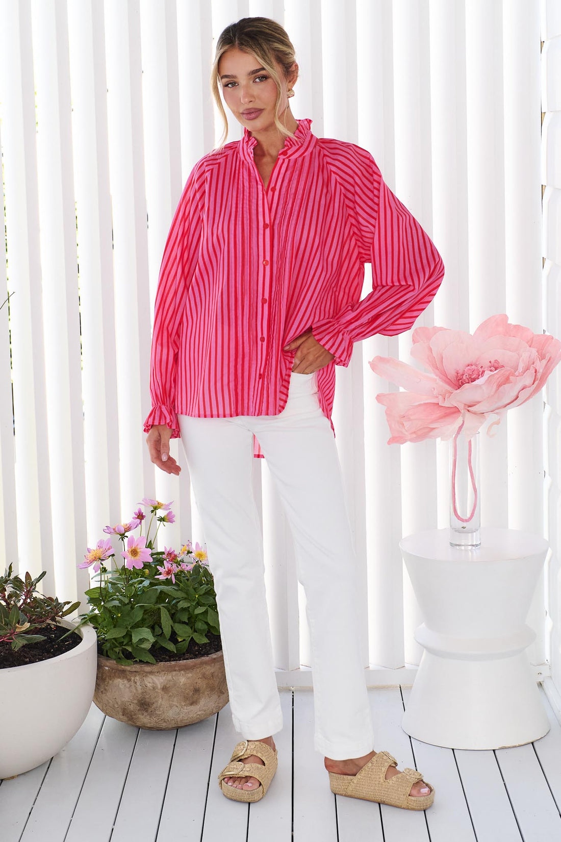 A woman wearing the vibrant pink and red stripe minnie + MAXXIE Cece Shirt with white pants, standing in front of a white panelled wall with potted flowers in the background.