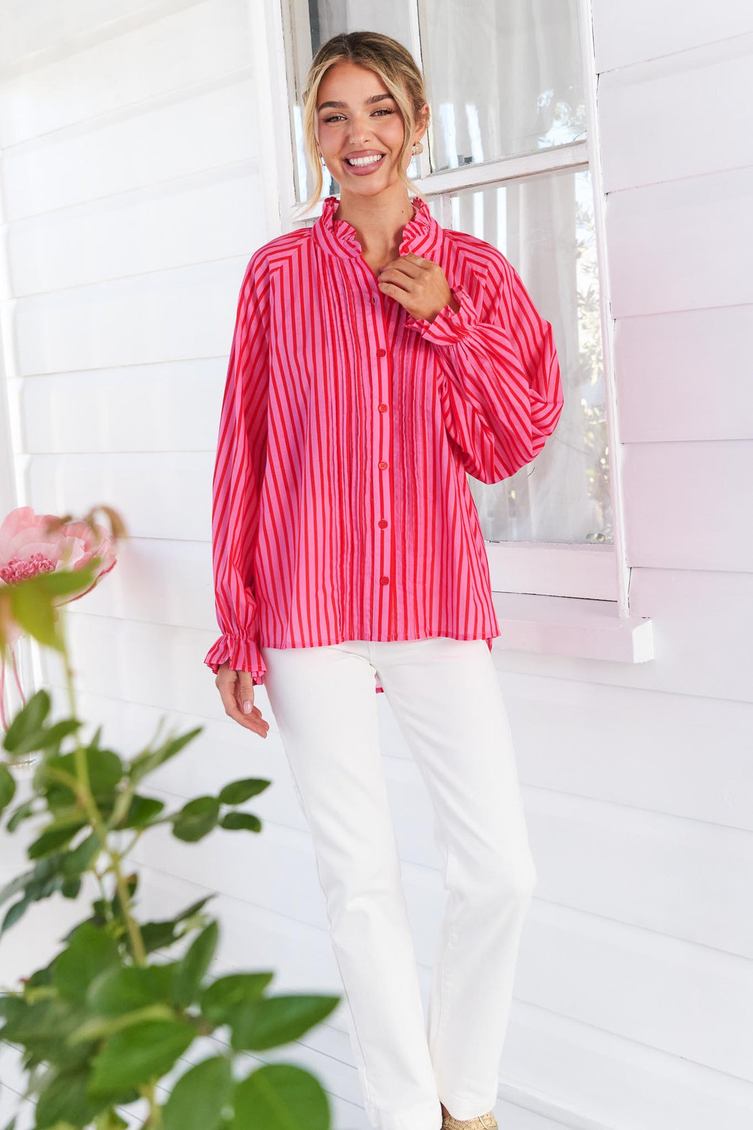 A woman wearing the vibrant pink and red stripe minnie + MAXXIE Cece Shirt with white pants, standing in front of a white house exterior with a window and green plants in the foreground.