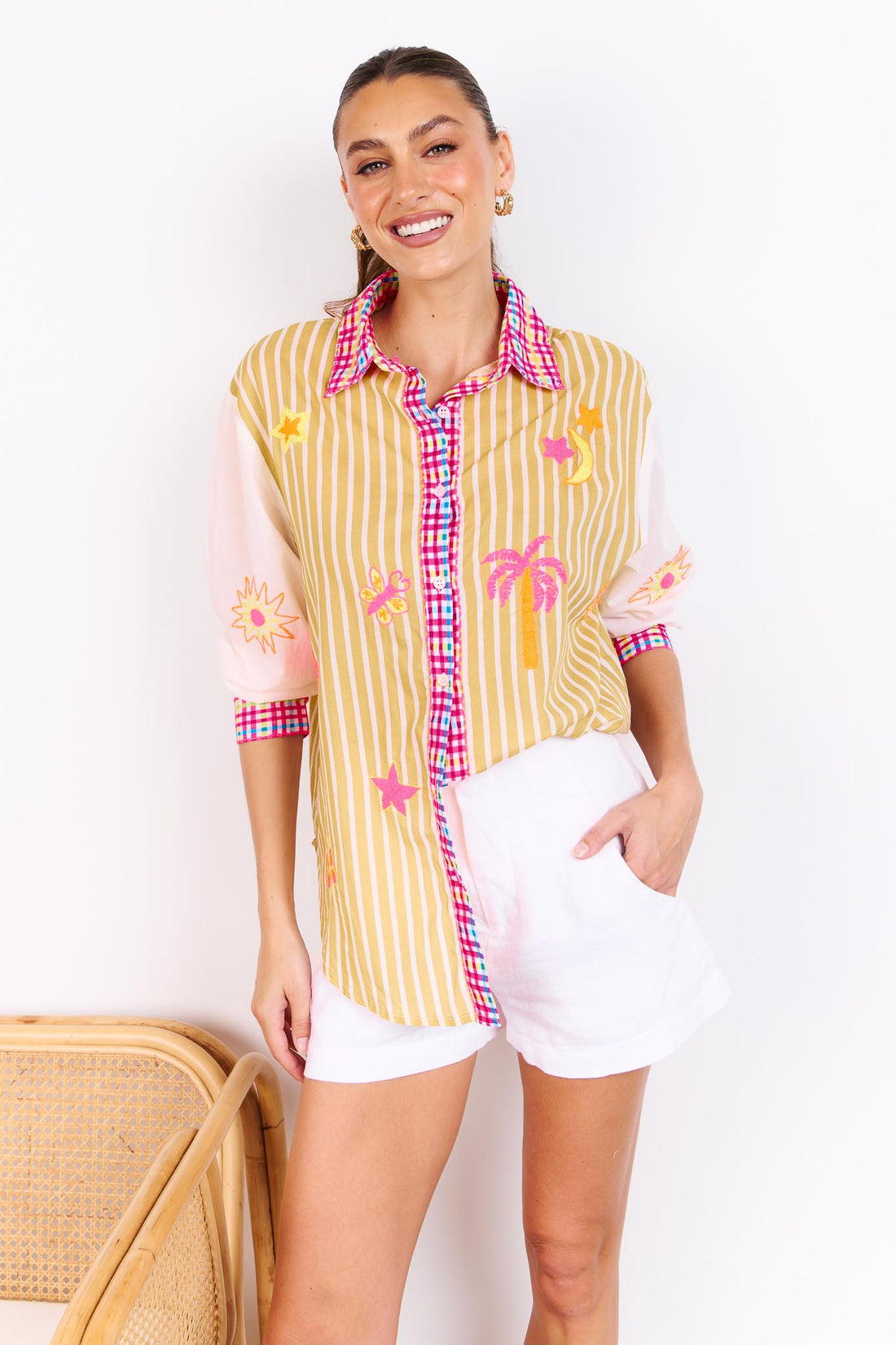Woman wearing the minnie + MAXXIE Ciao Bella Cotton Shirt with a tan and white stripe print, gingham trims and neon embroidered motifs paired with white shorts and standing in front of a white wall.