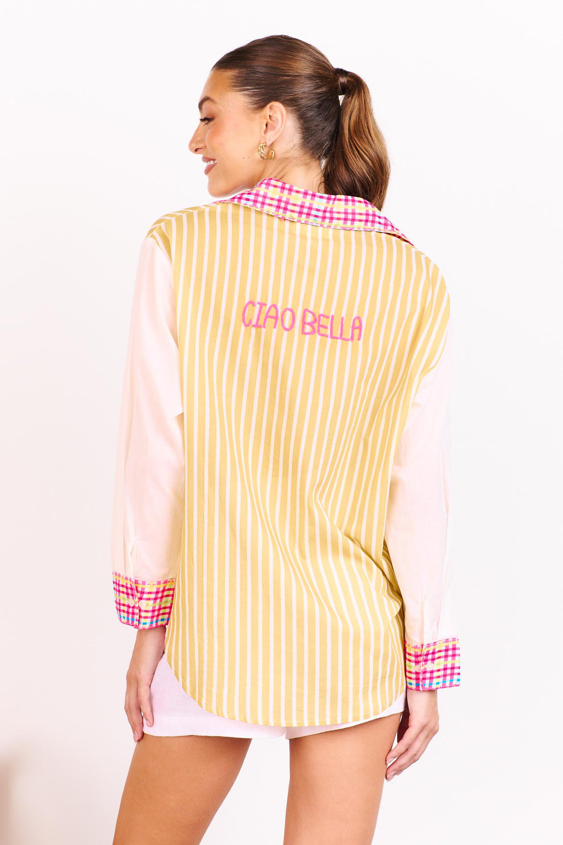 Back view of a woman wearing the minnie + MAXXIE Ciao Bella Cotton Shirt with a tan and white stripe print, gingham trims and a neon pink embroidered 'Ciao Bella', paired with white shorts and standing in front of a white wall.