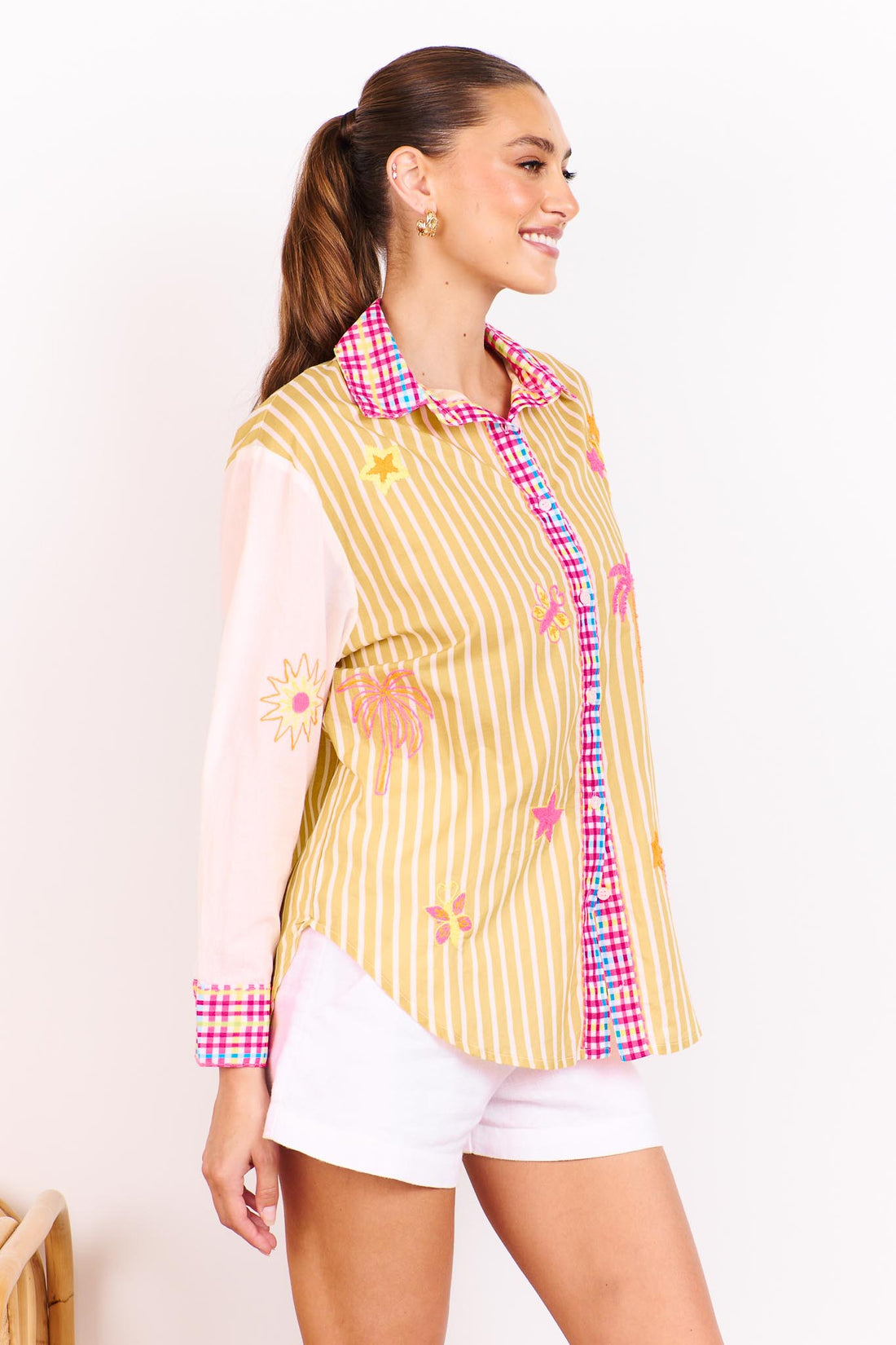 Woman wearing the minnie + MAXXIE Ciao Bella Cotton Shirt with a tan and white stripe print, gingham trims and neon embroidered motifs paired with white shorts and standing in front of a white wall.