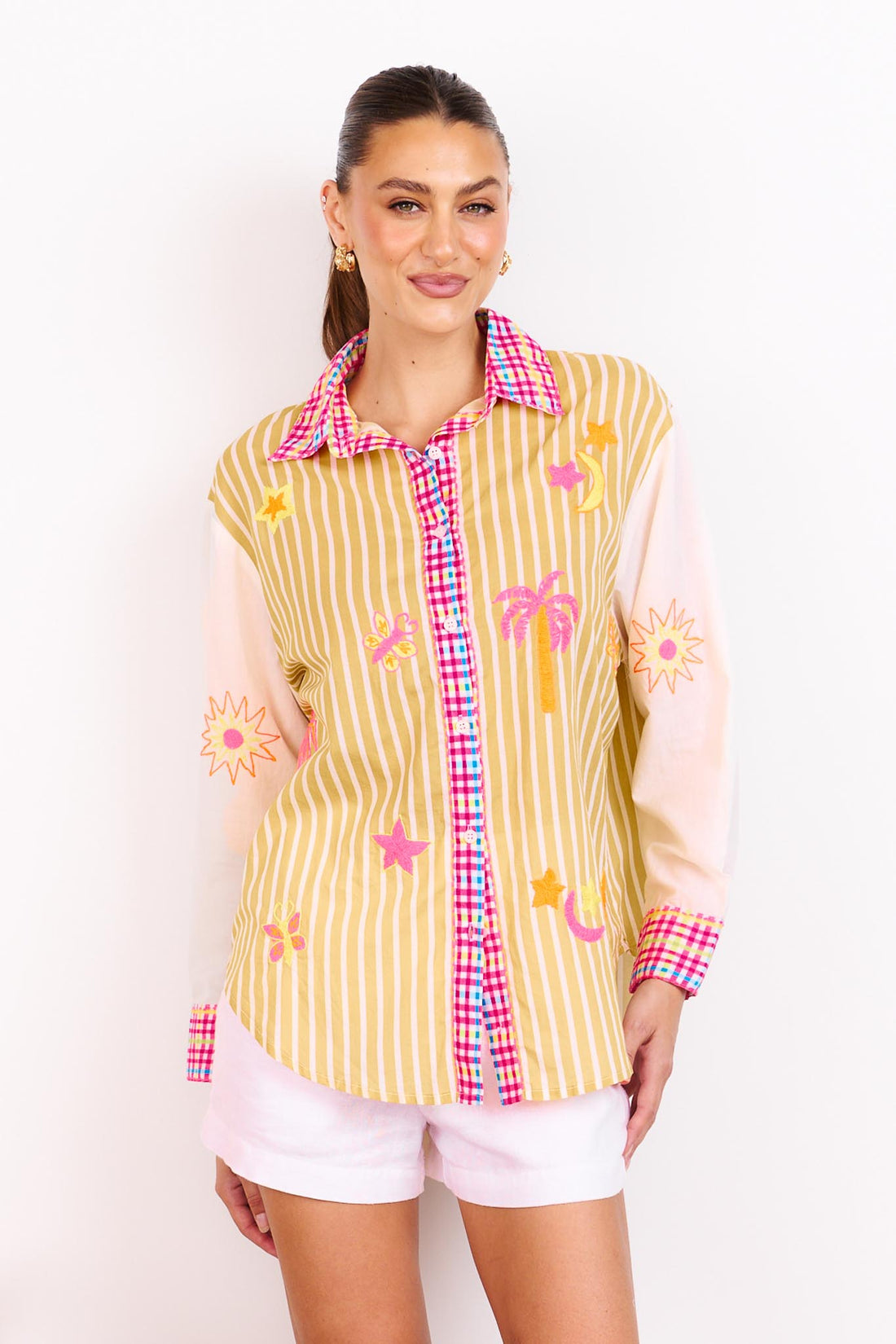 Woman wearing the minnie + MAXXIE Ciao Bella Cotton Shirt with a tan and white stripe print, gingham trims and neon embroidered motifs paired with white shorts and standing in front of a white wall.