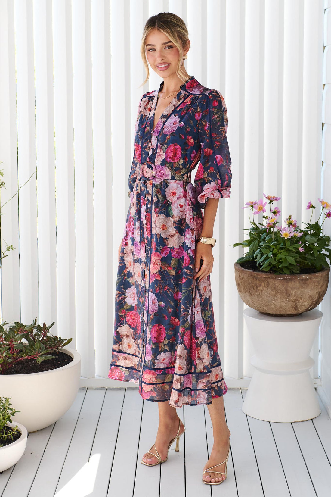 A woman wearing the minnie + MAXXIE Ciara navy blue midi shirt dress with a pink, red, and white floral print, standing on a white deck with potted plants in the background.