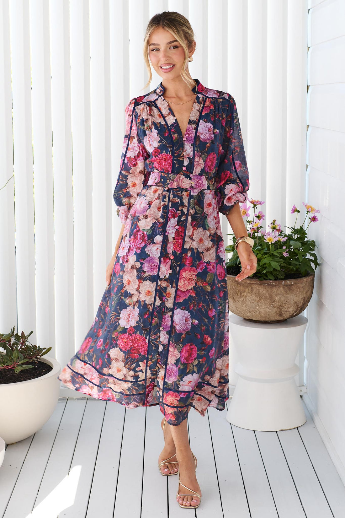 A woman wearing the minnie + MAXXIE Ciara navy blue midi shirt dress with a pink, red, and white floral print, standing on a white deck with potted plants in the background.